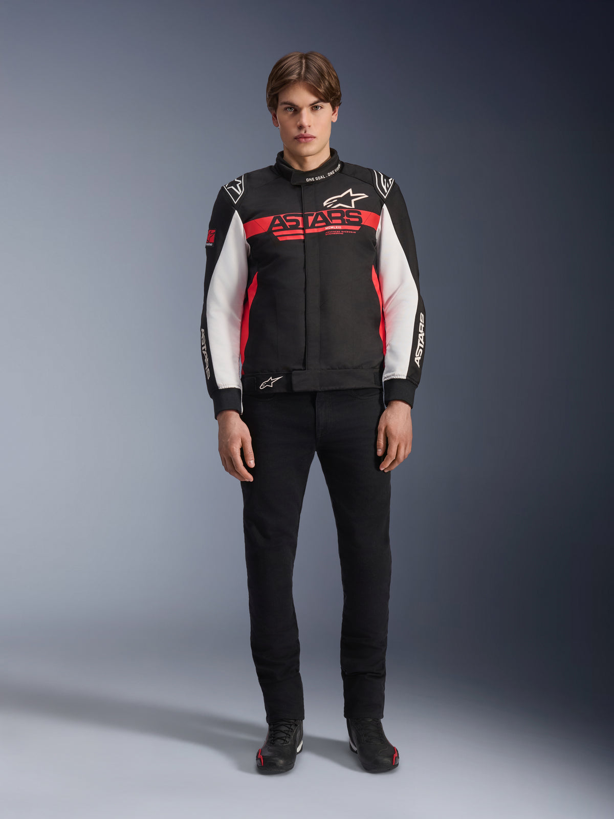 Männliches Modell in der Alpinestars Monza Sport Jacke in Schwarz, Leuchtend Rot und Weiß, mit einem großen roten „ASTARS“-Logo auf der Brust und weißen Einsätzen an den Ärmeln, kombiniert mit einer schwarzen Hose und schwarzen Motorradstiefeln.