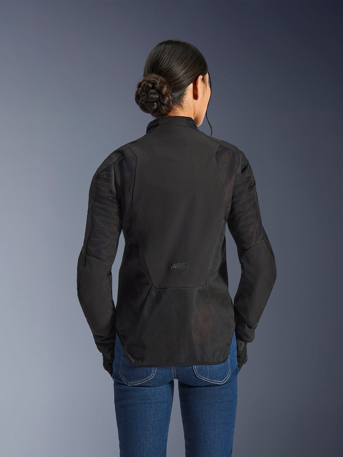 Alpinestars Woman Stella Aeroshell Airflow Jacket, Black Anthracite, rear view showing integrated mesh ventilation panels on the back and sleeves, laser-perforated softshell chassis with an elongated rear profile, worn with blue denim jeans.