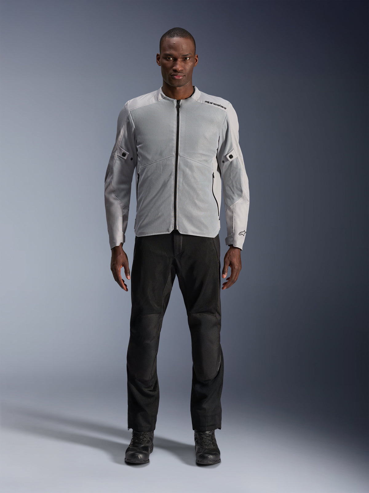 Alpinestars C-1 Air Jacke in Silber, urbane Textil-Motorradjacke für Herren mit belüfteten Mesh-Einsätzen, getragen von einem Model mit schwarzer Motorradhose und schwarzen Stiefeln, moderner Pendler-Stil