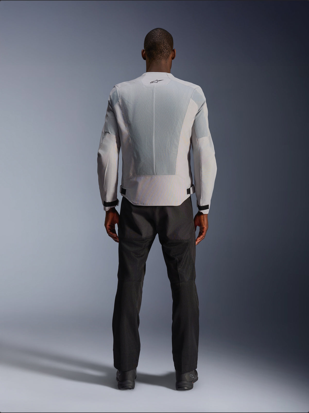 Alpinestars C-1 Air Jacke in Silber, Rückansicht an einem männlichen Model, Textil-Motorradjacke mit großen Tech-Mesh-Belüftungspanelen, kleinem schwarzen Logo im Nacken und Taillenverstellern, getragen mit einer schwarzen Motorradhose für einen modernen urbanen Look.