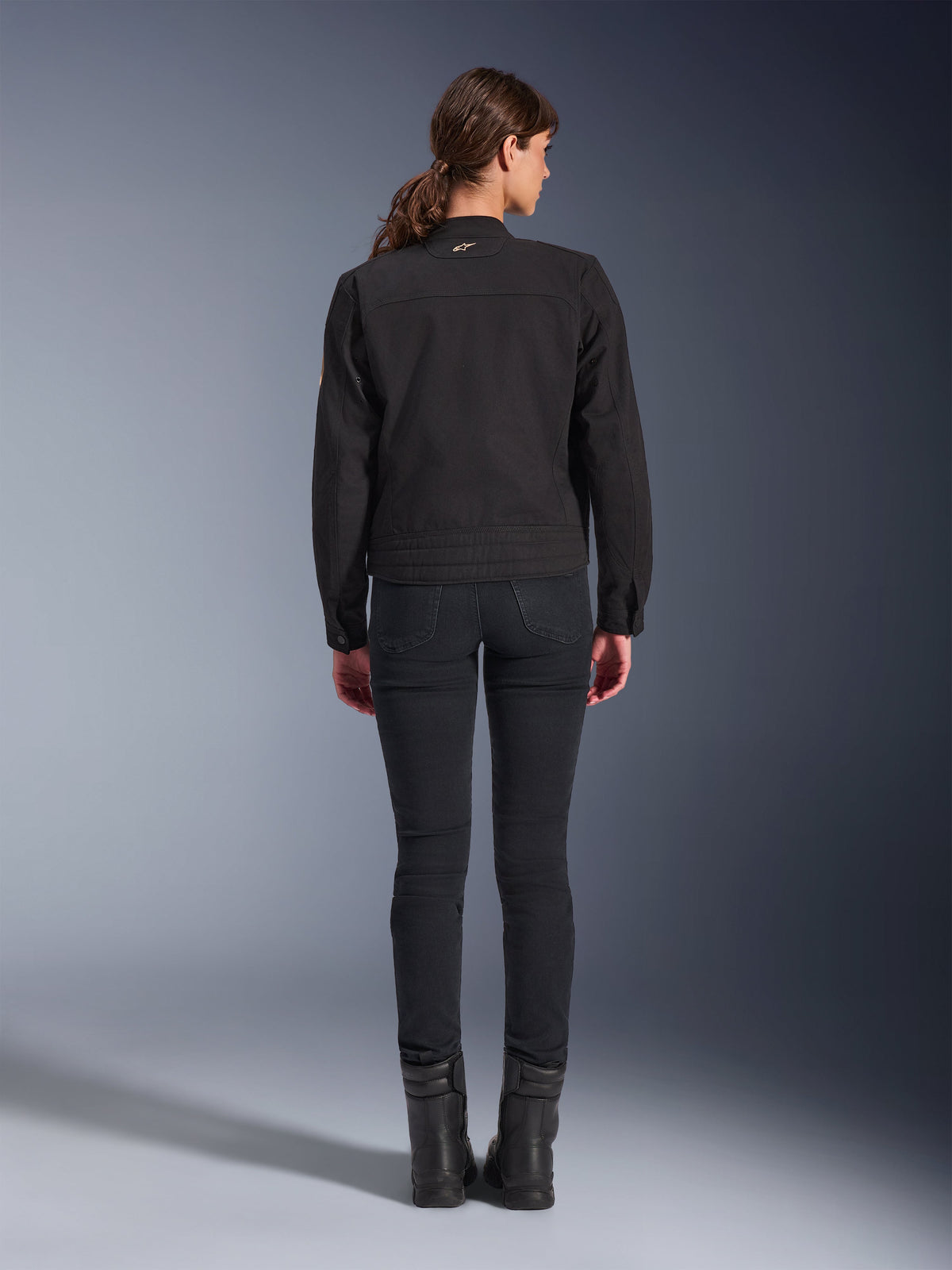 Alpinestars Stella Grace Jacke, Urbane Motorradjacke für Damen, Schwarz, Rückansicht zeigt Workwear-inspiriertes Design, getragen mit schwarzer Slim-Fit-Hose und schwarzen Stiefeln, Studioaufnahme