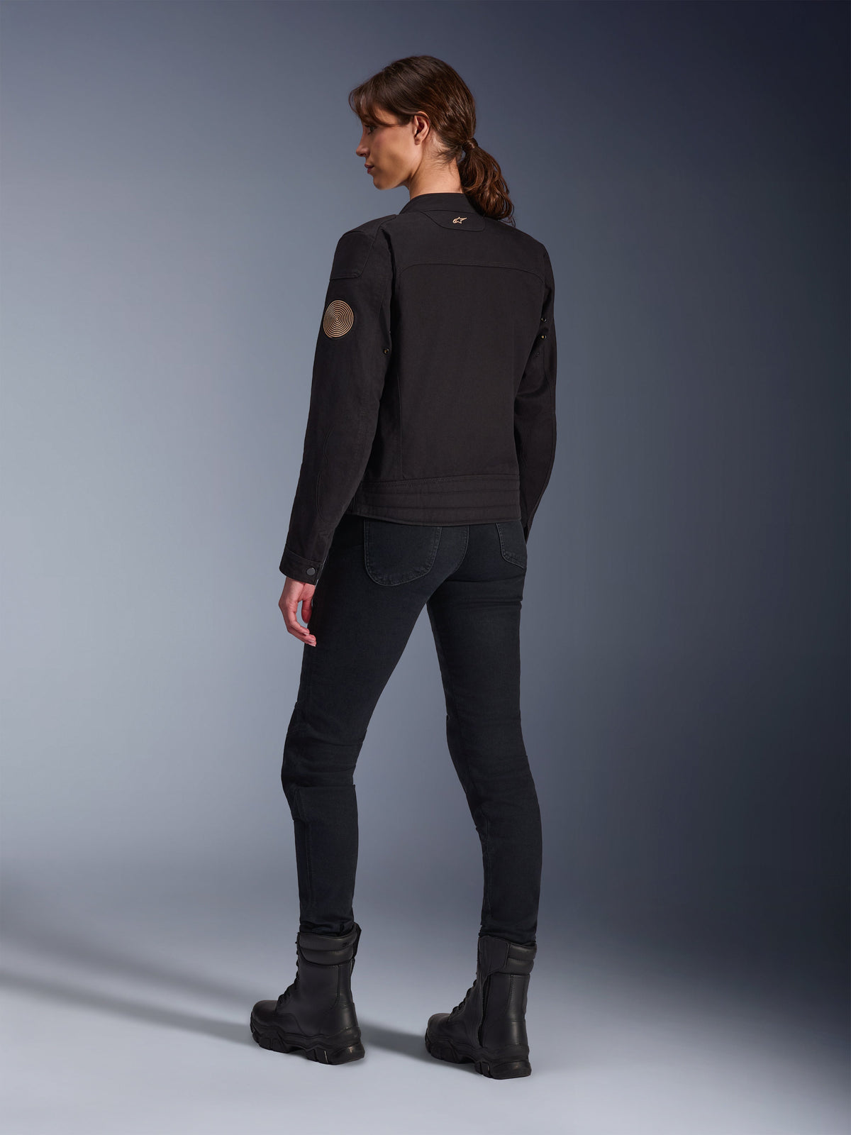 Model in der Alpinestars Stella Grace Jacke in Schwarz, Rückansicht mit Fokus auf den Vintage-Oscar-Spiralaufnäher am Arm und das kleine Stern-Logo am Kragen, kombiniert mit schwarzer Motorradhose und schwarzen Stiefeln für urbanes Fahren