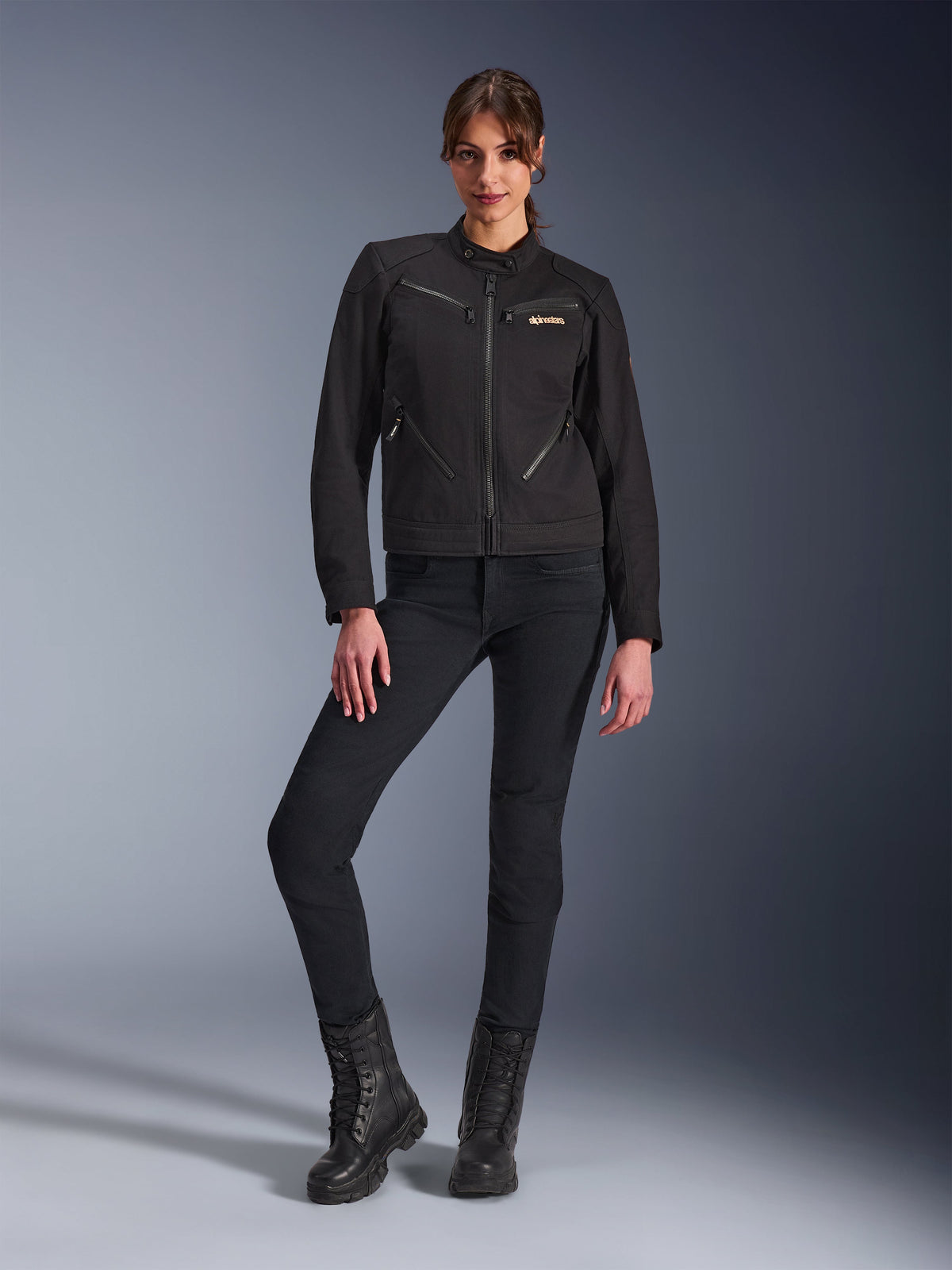 Frau in der Alpinestars Stella Grace Jacke in Schwarz, urbane Motorradjacke mit Workwear-Ästhetik, mit Reißverschlusstaschen vorne und Logo auf der Brust, kombiniert mit schmal geschnittener schwarzer Hose und schwarzen Stiefeln, moderne Motorradausrüstung für Damen.