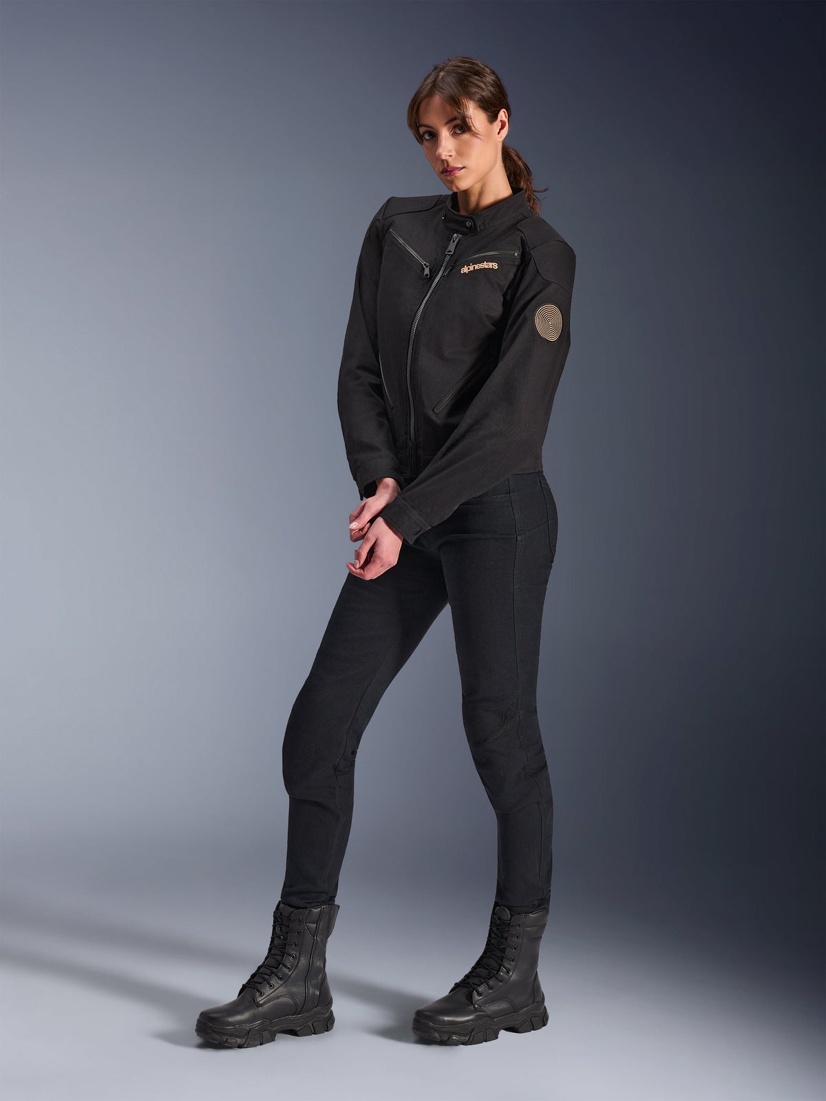Weibliches Model in der Alpinestars Stella Grace Damenjacke, schwarze Urban-Motorradjacke, kombiniert mit schwarzer Motorradhose und Stiefeln, Workwear-inspiriertes Design mit bronzefarbenem gesticktem Logo auf der Brust und kreisförmigem Spiral-Aufnäher am Ärmel