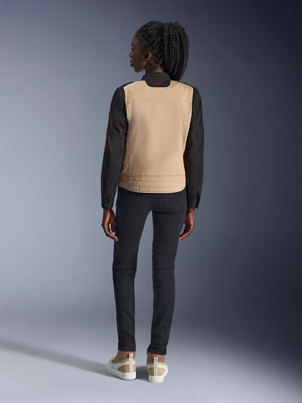 Woman wearing Alpinestars Stella Grace Jacket, Aluminum Black with tan torso and black sleeves, rear view, styled with black slim-fit pants and casual sneakers, urban motorcycle gear for women