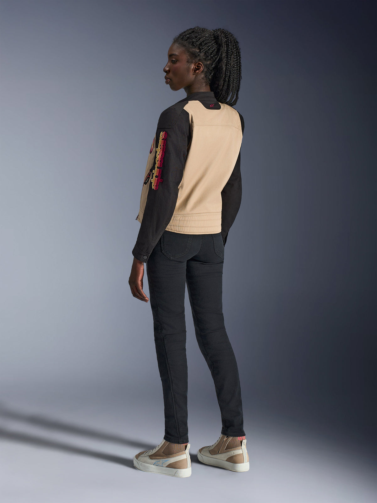 Alpinestars Woman Stella Grace Jacket, Urban Motorcycle Jacket, Aluminum Black with tan back panel and black sleeves, back-side view on a female model, featuring embroidered colorful branding on the sleeve and pleated shoulder construction.