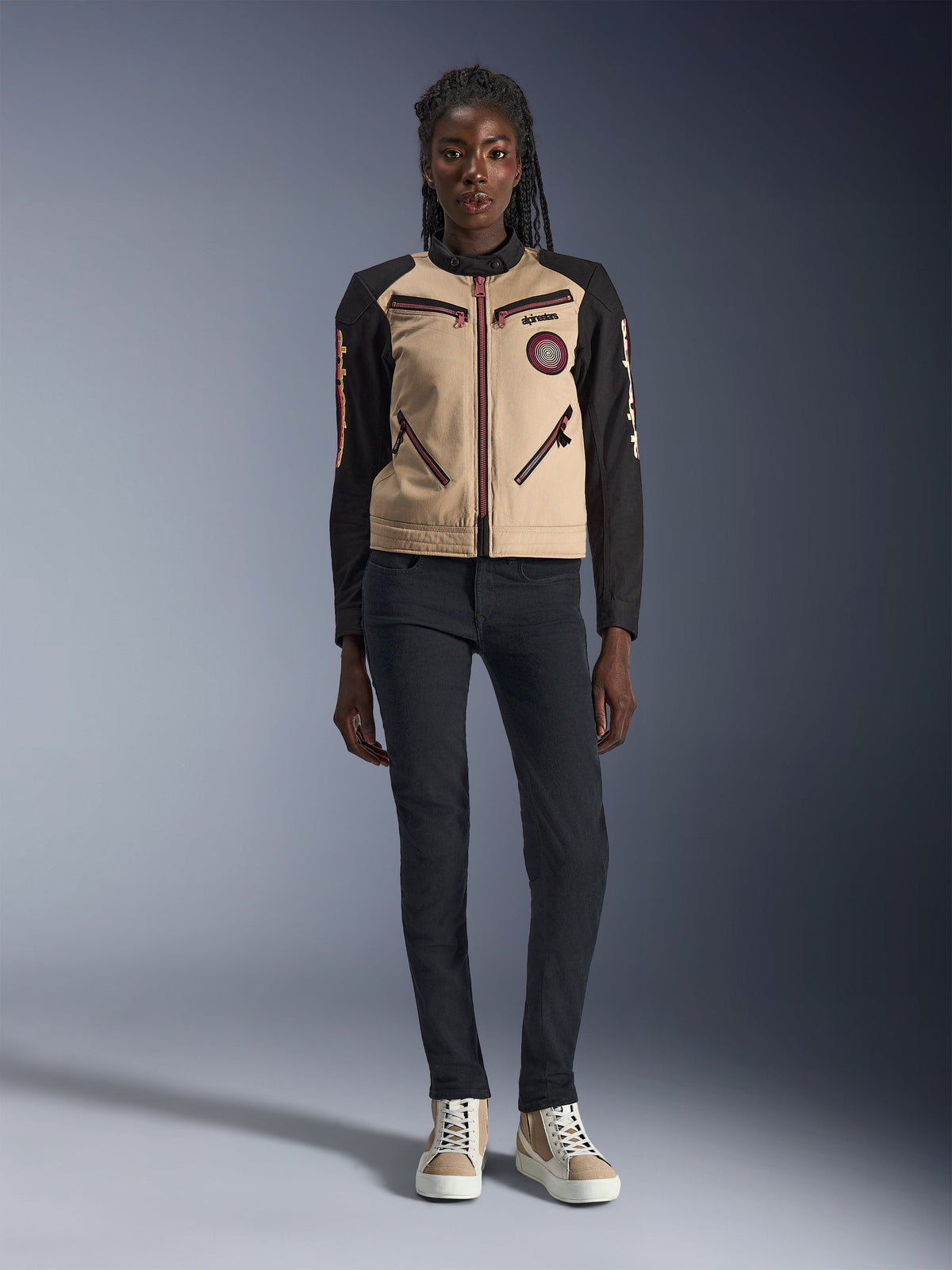 Alpinestars Woman Stella Grace Jacket, urban motorcycle jacket, tan and black textile construction, featuring front zippers and embroidered Oscar spiral patch, worn with black pants and high-top sneakers, contemporary workwear-inspired riding gear