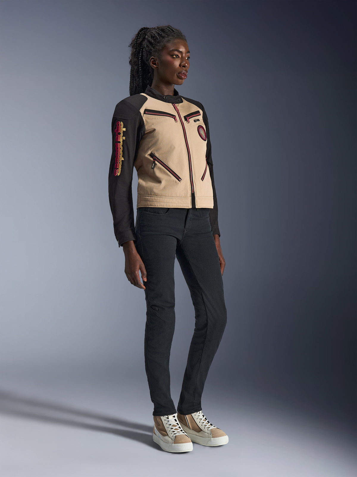 Model wearing Alpinestars Stella Grace Jacket in Aluminum Black, featuring a tan torso and black sleeves with red zipper accents, paired with slim-fit black pants and cream sneakers, urban contemporary motorcycle apparel for women
