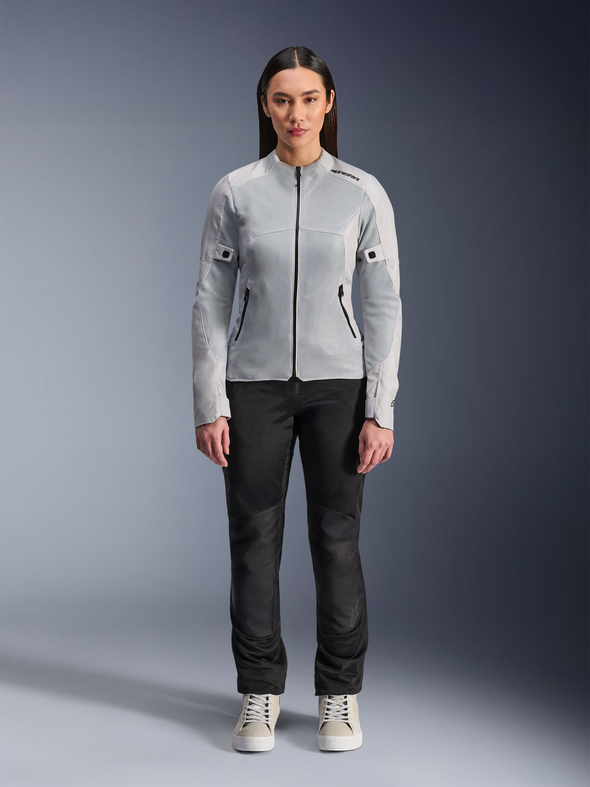 Frau in Alpinestars Stella C-1 Air Jacke in Silber, Vorderansicht, urbane Motorrad-Textiljacke mit Tech-Mesh-Einsätzen, kombiniert mit schwarzer Hose und weißen Sneakern, moderne Motorradausrüstung