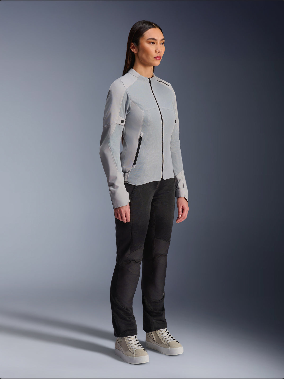 Frau in der Alpinestars Stella C-1 Air Jacke in Silber, leichte Textil- und Mesh-Motorradjacke, kombiniert mit einer schwarzen Fahrhose und lässigen Sneakern, schlichte urbane Riding-Ästhetik.
