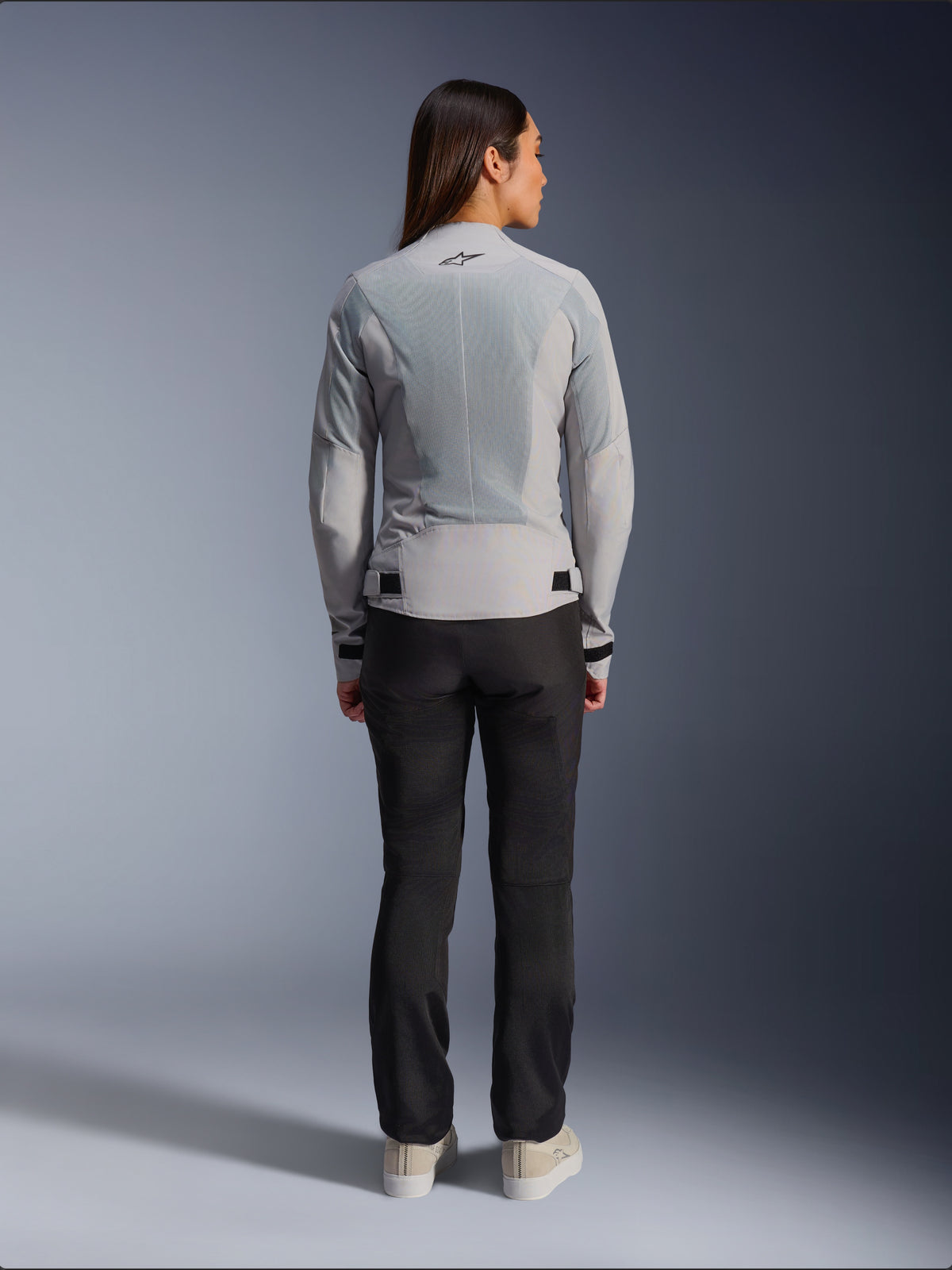 Frau in der Alpinestars Stella C-1 Air Jacke in Silber, Rückansicht, große Tech-Mesh-Einsätze zur Belüftung, reflektierendes Stern-Logo, kombiniert mit einer schwarzen Motorradhose und weißen Sneakern, moderne urbane Motorradausrüstung
