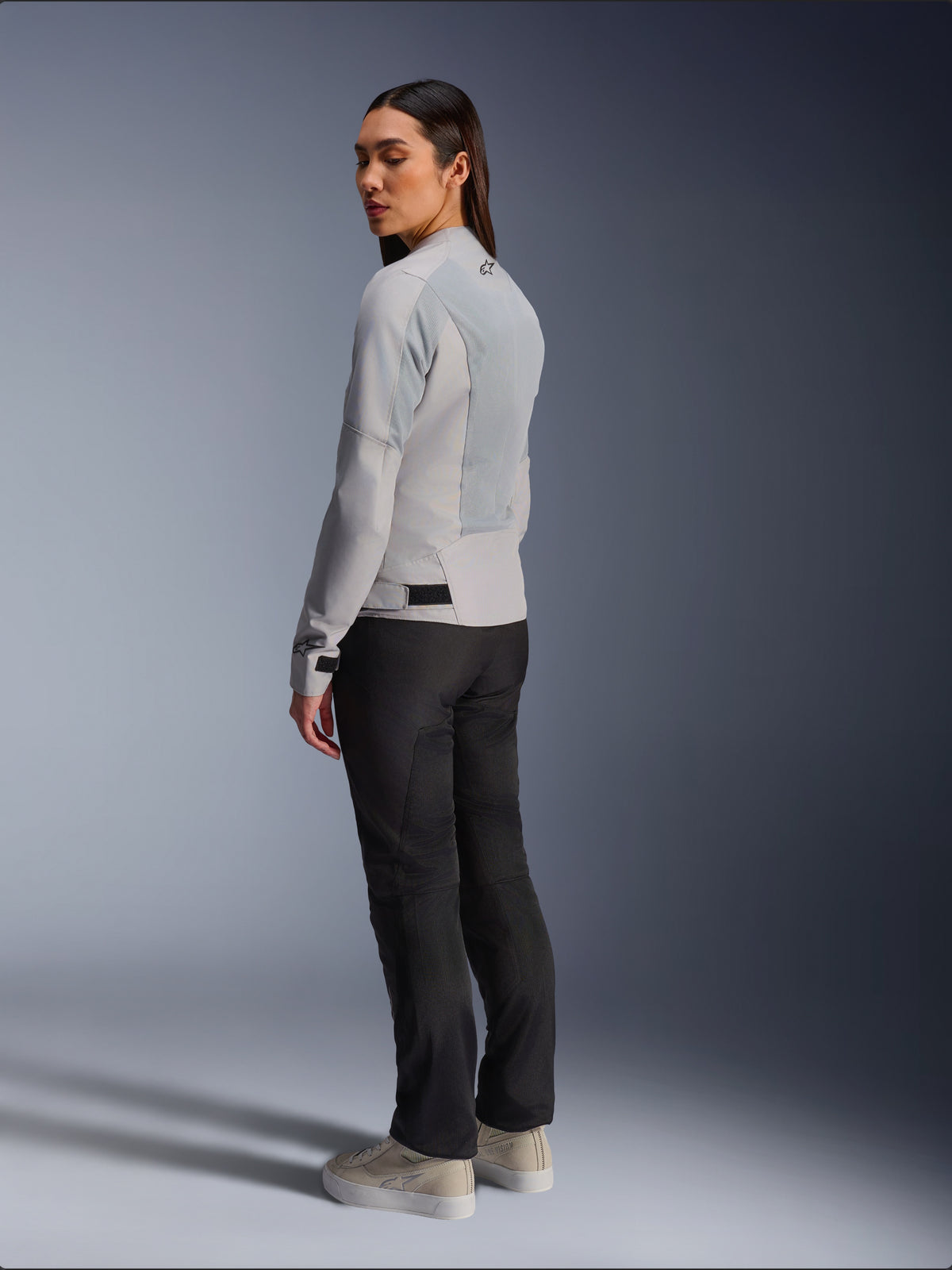 Frau trägt Alpinestars Stella C-1 Air Jacke in Silber, Rück- und Seitenansicht mit Tech-Mesh-Einsätzen und reflektierendem Schulter-Logo, kombiniert mit schwarzer Motorradhose und beigen Sneakern, moderne urbane Fahr-Ästhetik