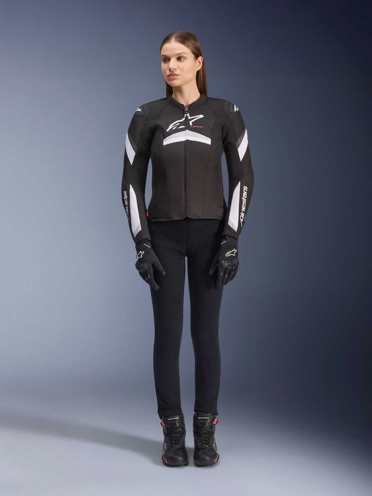 Female model wearing the Alpinestars Stella T-GP Plus R V4 Jacket in Black and White, textile sport motorcycle jacket, paired with black riding pants, gloves, and boots, front view showing white Alpinestars logo and sleeve accents.