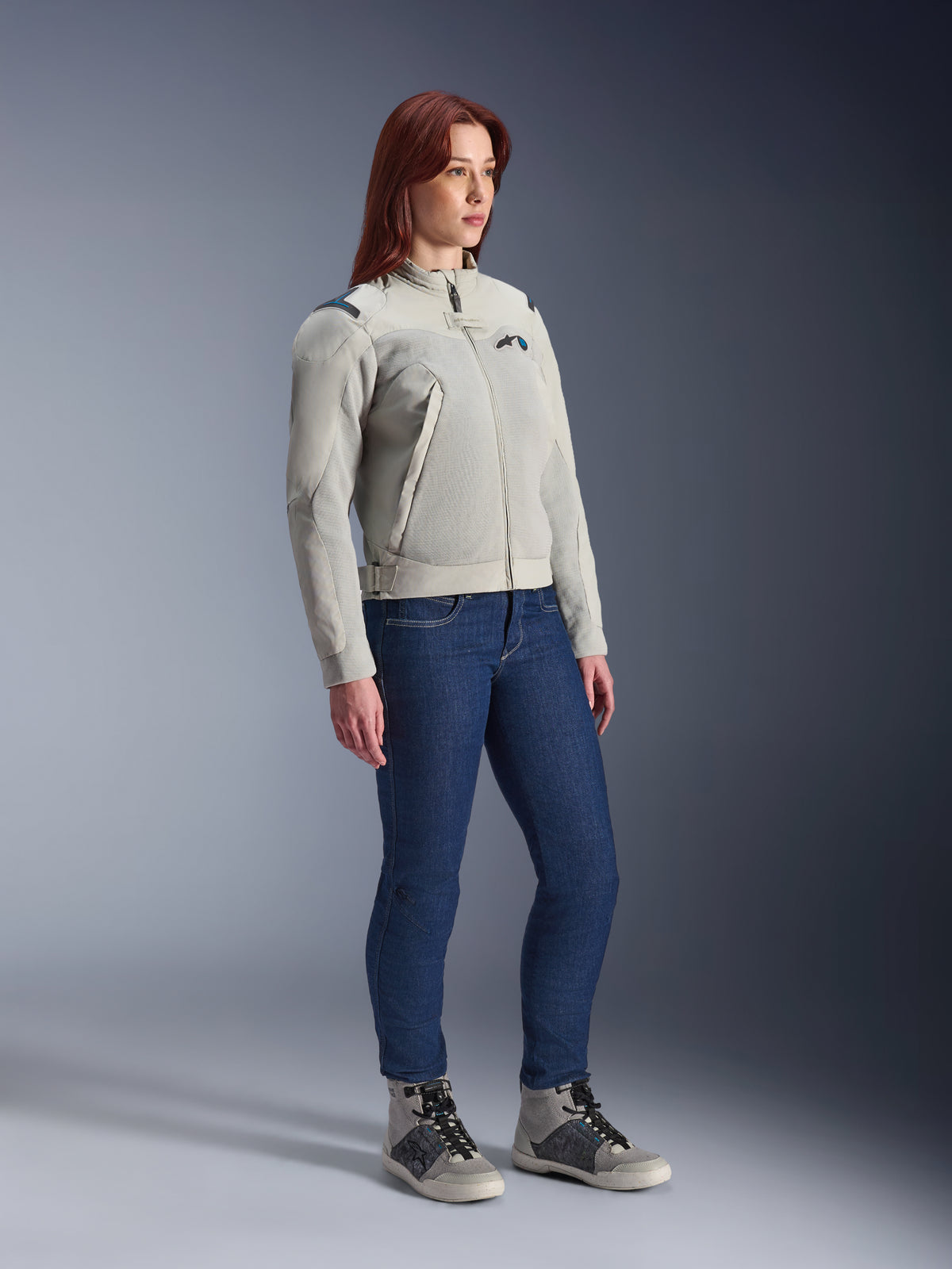 Frau trägt die Alpinestars Stella Circal-1 Air Jacke in Metal Gray und Methyl Blue, kombiniert mit blauen Jeans und lässigen Motorradschuhen, zeigt die Mesh-Einsätze und das minimalistische urbane Design der Jacke.