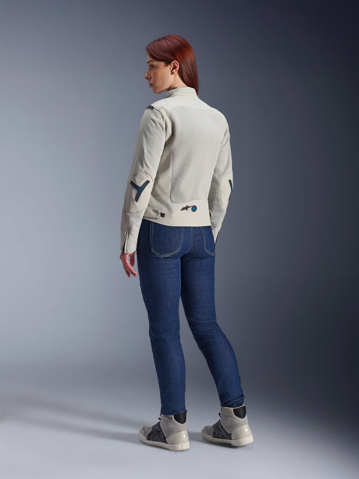 Frau in Alpinestars Stella Circal-1 Air Jacke in Metal Gray und Methyl Blue, Rückansicht mit Mesh-Belüftungseinsätzen und neonblauen Akzenten, kombiniert mit dunkelblauer Riding-Jeans und grauen Sneakern, urbaner Motorrad-Lifestyle-Shot.