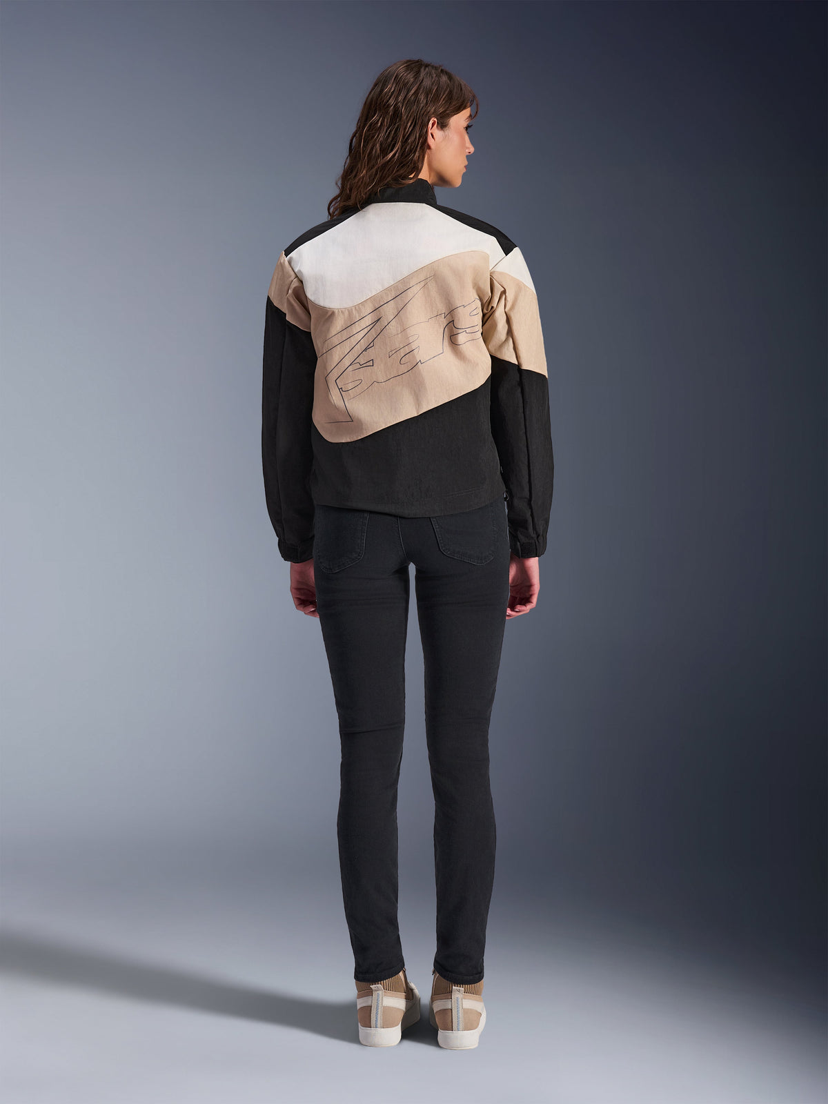 Alpinestars Stella Moshell Jacke, urbane Motorradjacke für Damen, Rückansicht, asymmetrisches Design in Black Sand und Off White mit großem grafischem Umriss, getragen mit schwarzer Slim-Fit-Hose und beigen Sneakern, urbane Fahrausrüstung