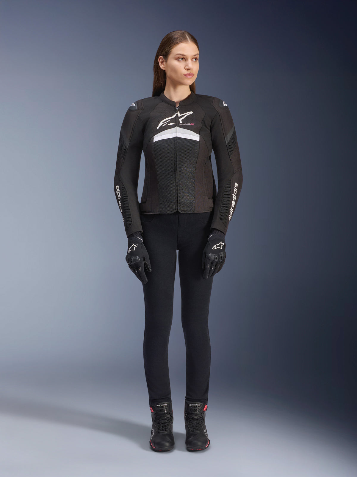 Female rider wearing the Stella T-GP Plus R V4 Airflow Jacket, Black and White, paired with black riding pants, gloves, and boots, featuring mesh panels for ventilation and white Alpinestars branding on the chest and arms