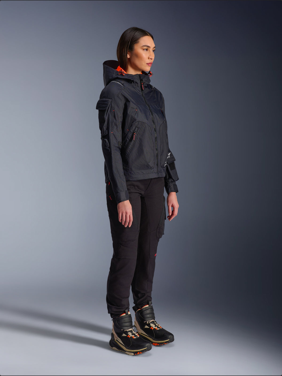Motorradfahrerin in der Alpinestars Molly Air Jacke in Schwarz Fluo Coral, leichte Textil-Motorradjacke mit leuchtend orangefarbenem Kapuzenfutter, kombiniert mit schwarzer Motorradhose und technischen Stiefeln, urbanes zeitgenössisches Utility-inspiriertes Design.