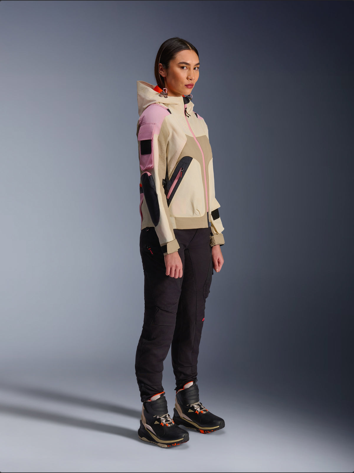 Weibliches Modell in der Alpinestars Molly Air Jacke in Sand Pink Almond, kombiniert mit dunkler technischer Hose und hohen Fahrstiefeln, urbane Motorradjacke mit Kapuze und mehrfarbigen Einsätzen, zeitgenössisches, von Streetwear inspiriertes Design