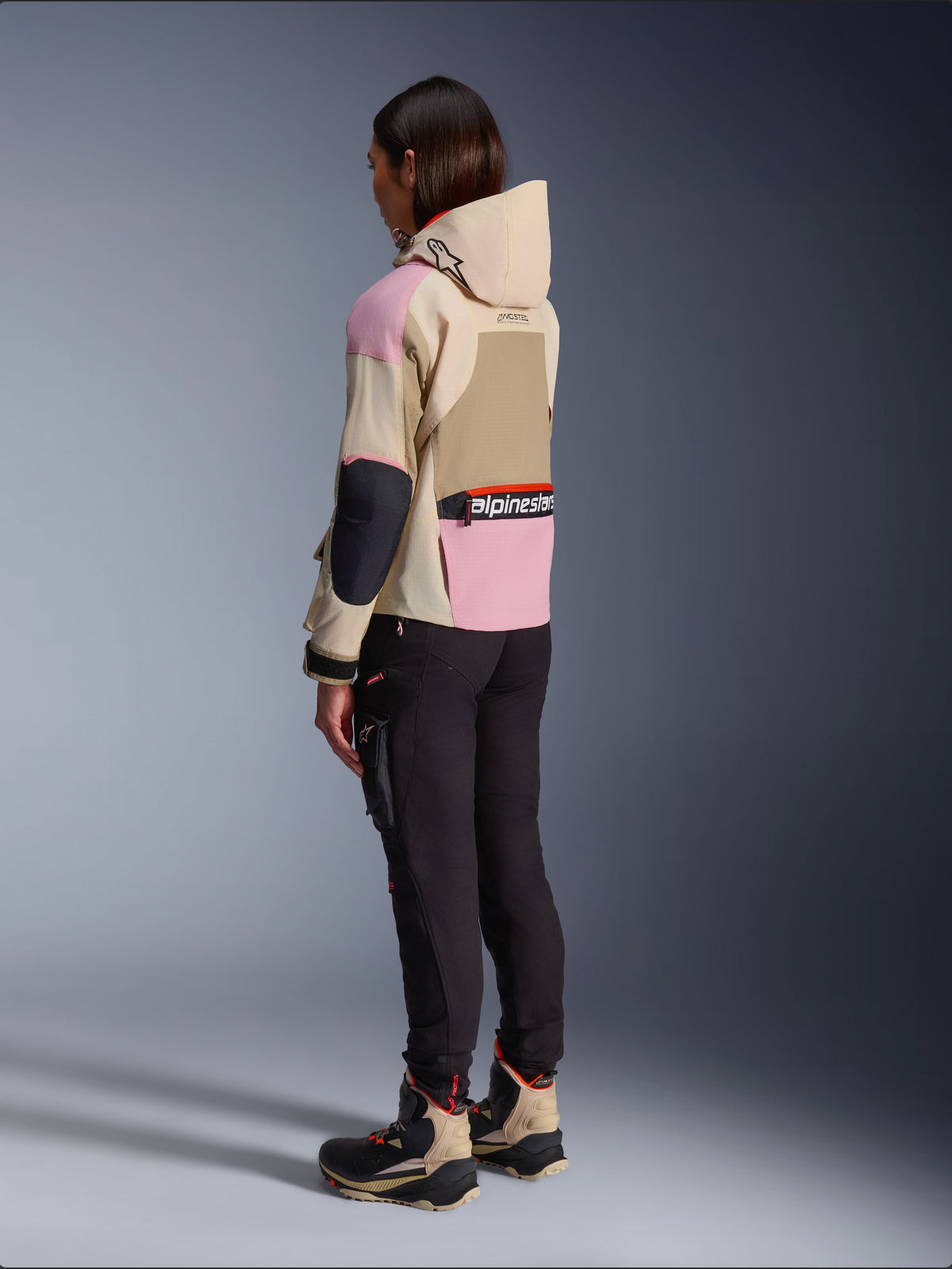 Motorradfahrerin in der Alpinestars Molly Air Jacke in Sand Pink Almond, mehrfarbiges Design in Beige und Rosa, seitliche Rückansicht mit schwarzer Motorradhose und passenden technischen Stiefeln, urbaner Motorradstil mit Kapuze.