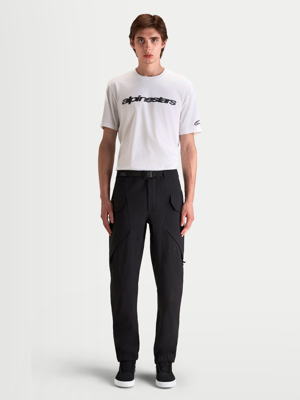 Alpinestars Juggernaut V 2 Drystar Hose, Herren-Urban-Motorradhose in Schwarz, Vorderansicht an einem Model mit weißem T-Shirt und lässigen Motorradschuhen, zeigt Cargotaschen und einen verstellbaren Taillengürtel