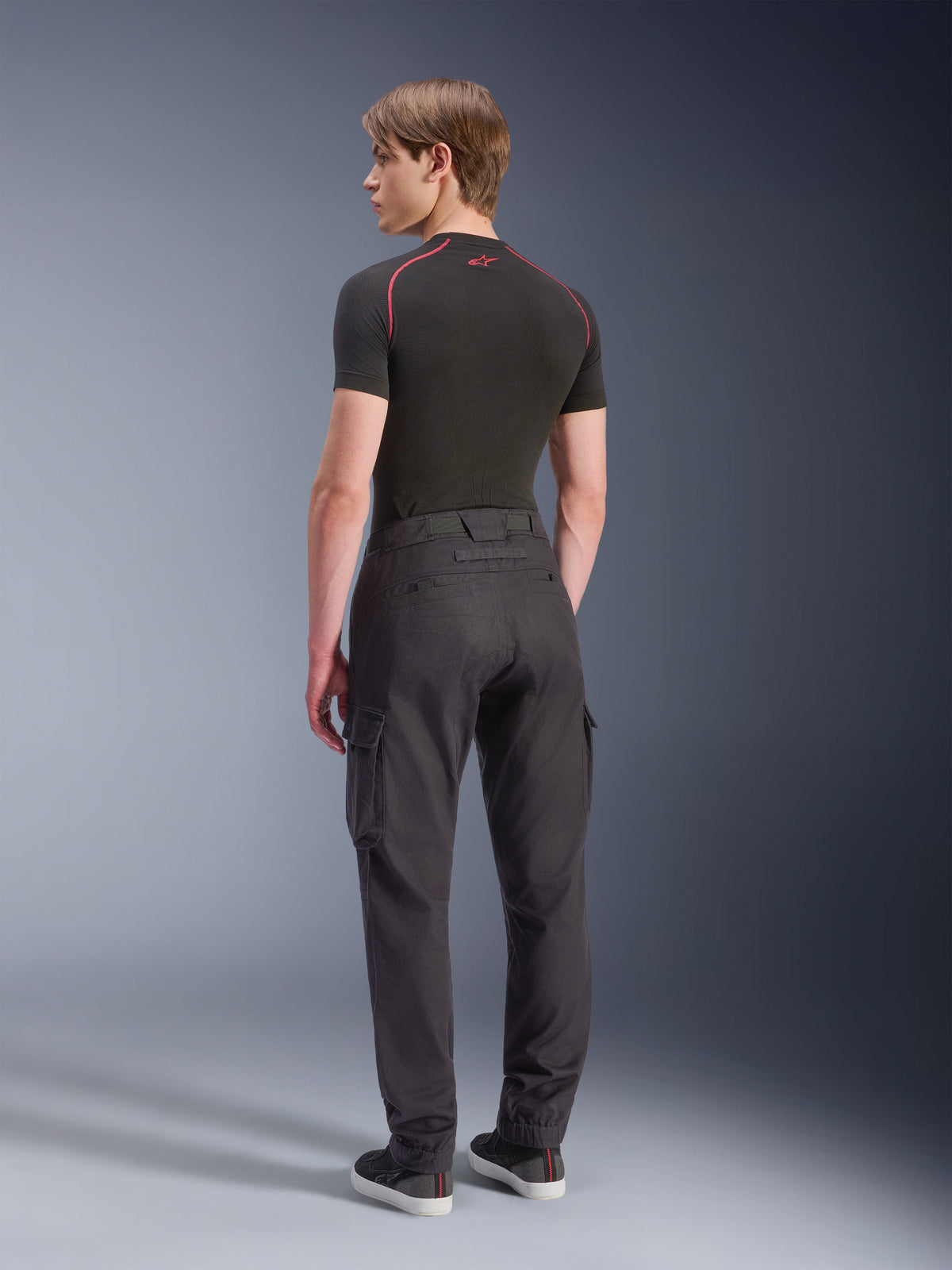 Alpinestars Flex-AST Cargo-Hose – Kurz, Motorrad-Cargo-Hose, Schwarz, Rückansicht eines Models in einer regulär geschnittenen, konisch zulaufenden Cargo-Hose mit Seitentaschen und elastischen Säumen, kombiniert mit einem schwarzen Baselayer und Freizeitschuhen