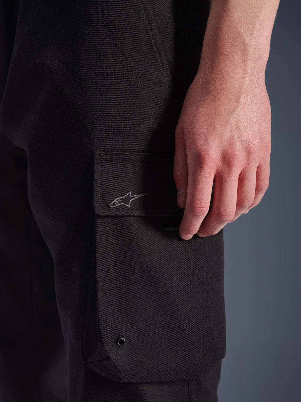 Alpinestars Flex-AST Cargo-Hose, Urbane Motorradhose, Schwarz, Nahaufnahme der seitlichen Cargotasche mit besticktem Astar-Logo, Metallöse und strapazierfähiger Stretch-Twill-Struktur