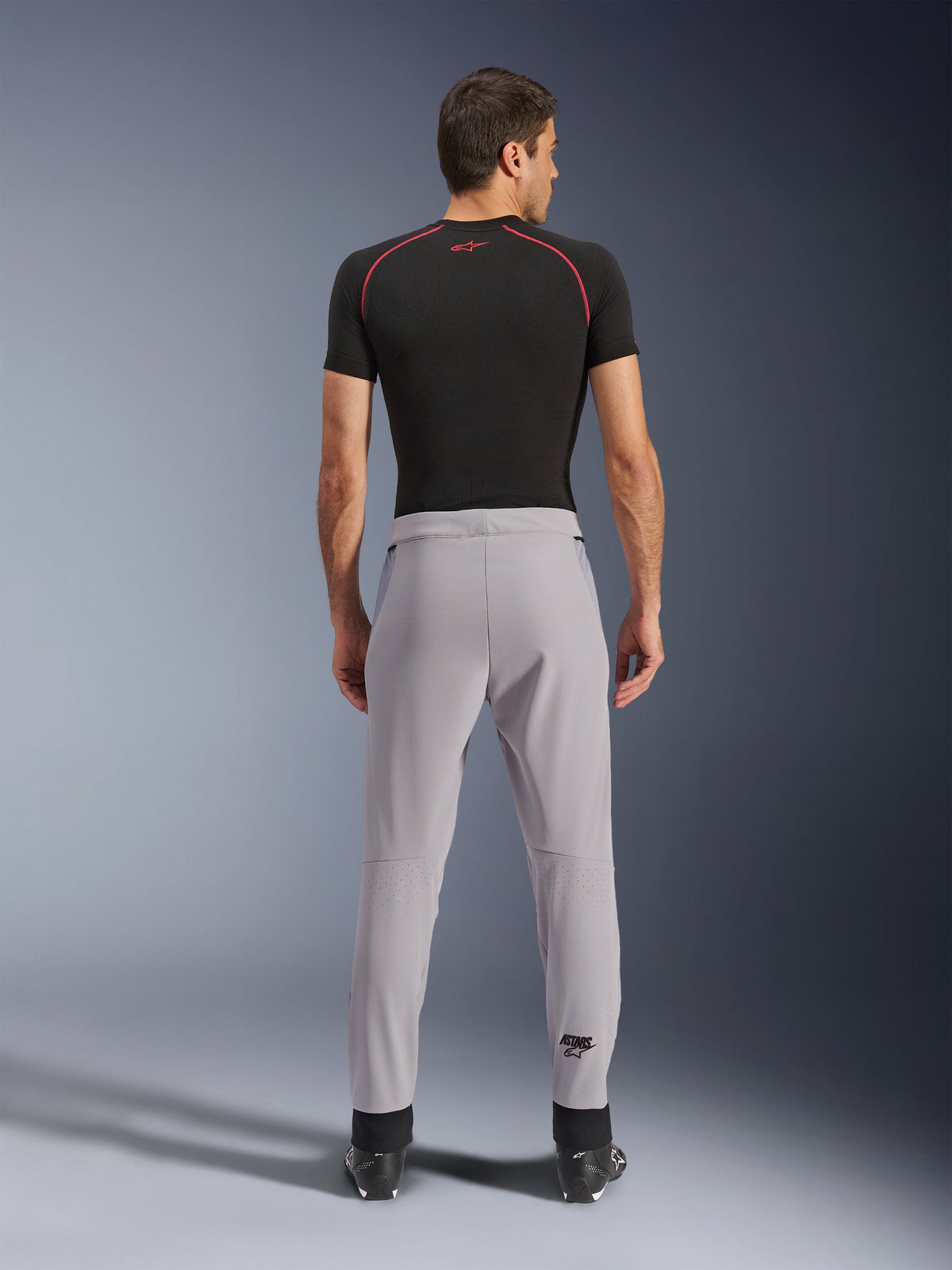Alpinestars Aeroshell Airflow Hose, Mittelgrau, Rückansicht mit laserperforierten Zonen an den Waden, leichte Stretch-Softshell-Konstruktion, kombiniert mit schwarzem Kurzarmshirt und Motorradstiefeln, Sport-Fit-Design für Fahrten bei warmem Wetter