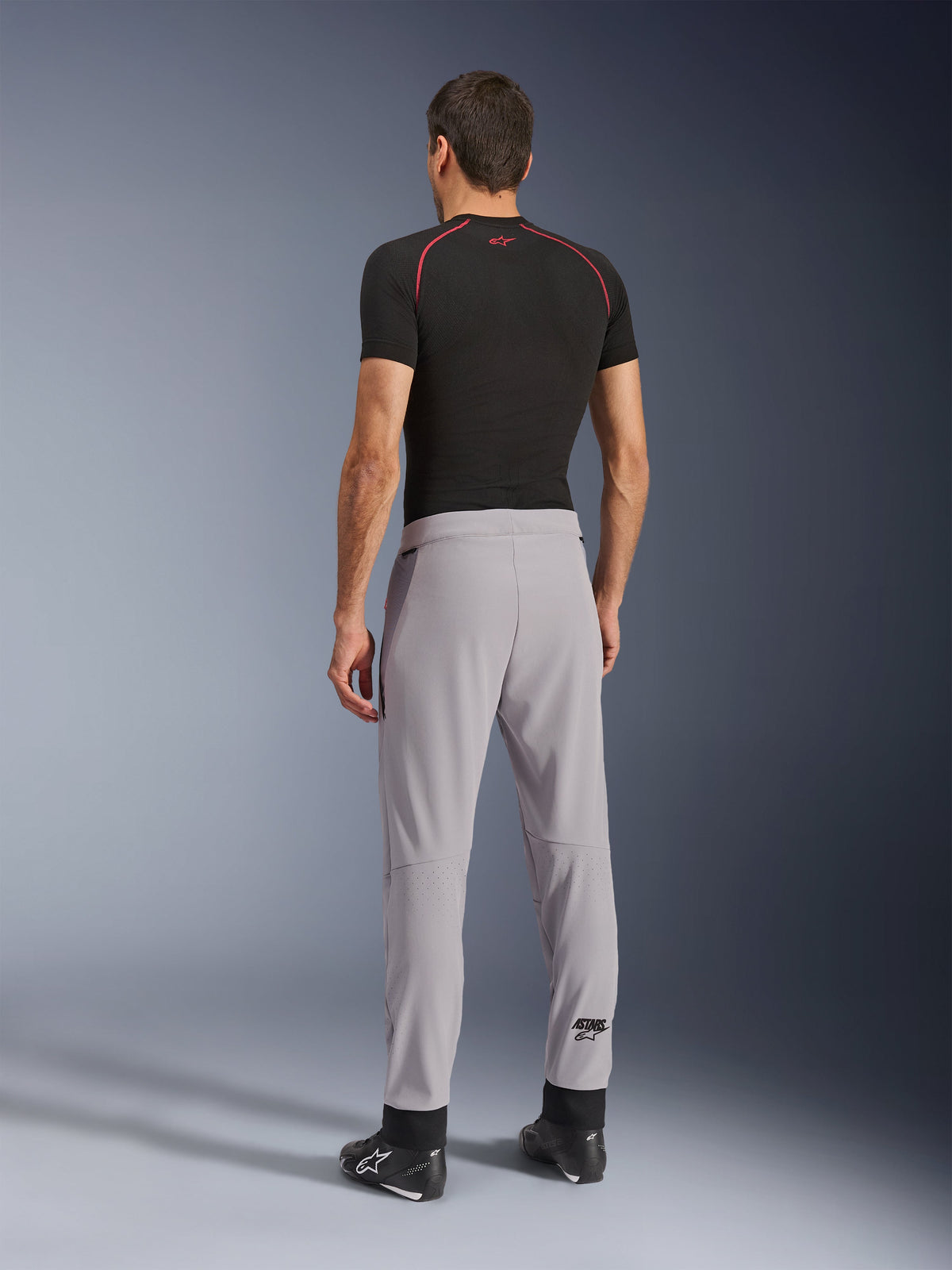 Rückansicht eines Modells in der Alpinestars Aeroshell Airflow Hose in Mid Gray, kombiniert mit einem schwarzen Base-Layer-Shirt und schwarzen Schuhen, wobei die laserperforierten Zonen an den Waden und die athletische Sportpassform hervorgehoben werden.