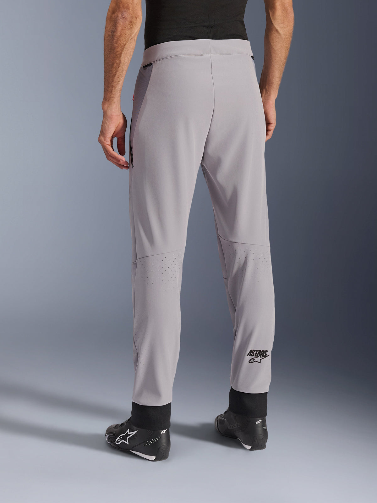 Alpinestars Aeroshell Airflow Hose, Mittelgrau, Rückansicht mit laserperforierten Zonen an den Waden, ASTARS-Logo am Unterschenkel, schwarze verstellbare Knöchelmanschetten, getragen mit schwarzen Motorradstiefeln, leichte Sport-Textil-Motorradhose