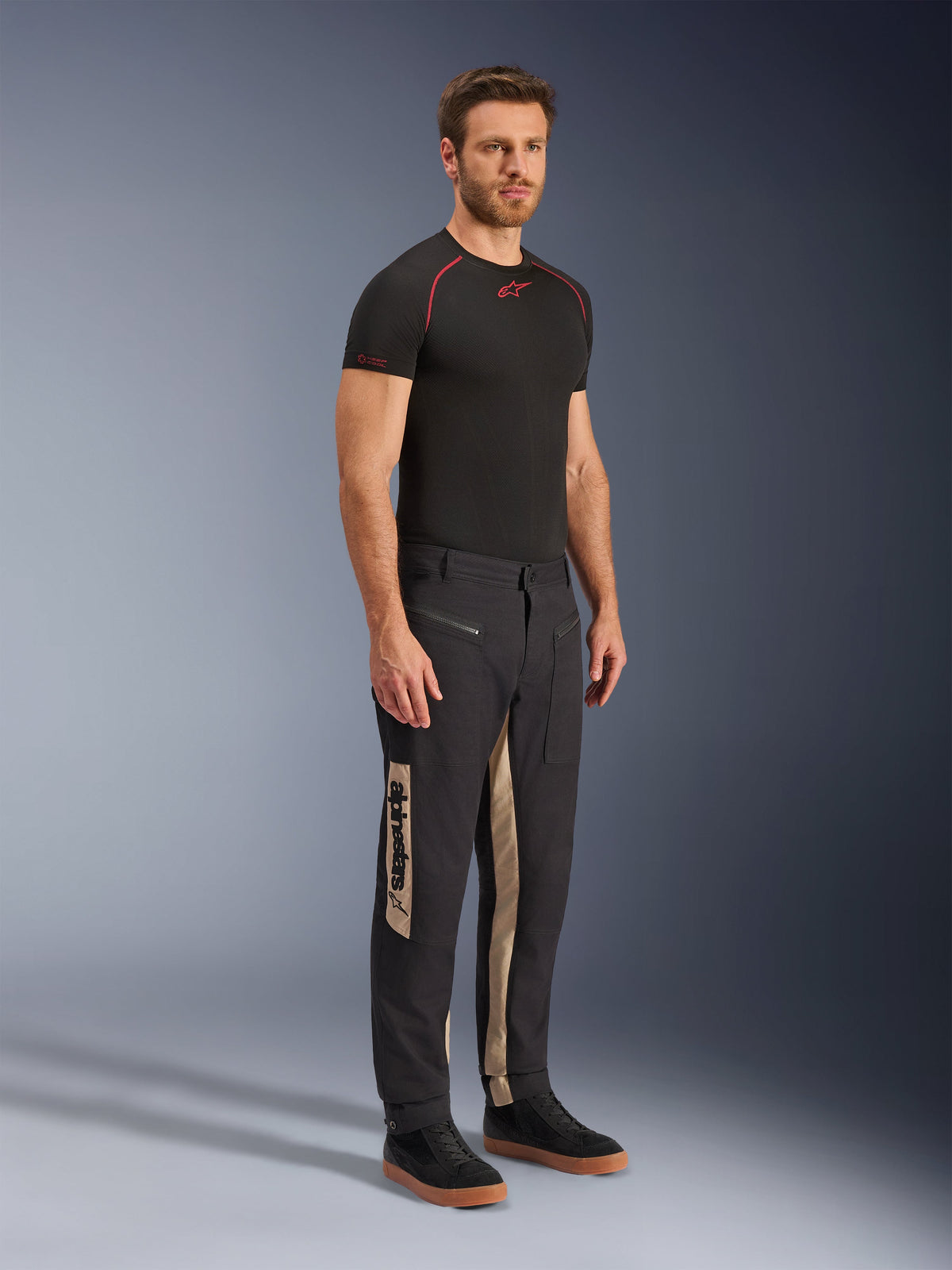 Alpinestars Flex-AST Canvas Pants, Urban Motorcycle Pants, Black Aluminum, worn with a black technical base layer and black sneakers, featuring tan side panels with vertical Alpinestars branding, utilitarian workwear-inspired design with pre-shaped knees and metal zippers
