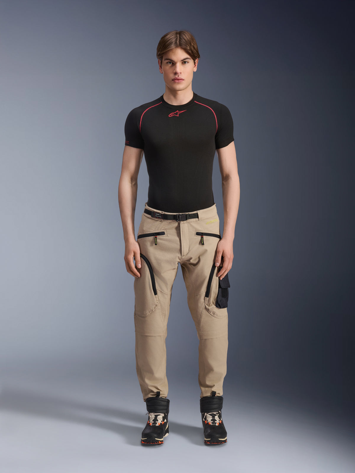 Männliches Model in Alpinestars Acteon Hose in Sand, kombiniert mit einem schwarzen kurzärmeligen Base Layer und schwarzen technischen Stiefeln, mit Cargotaschen mit schwarzen Reißverschlüssen und einer schmal zulaufenden Passform, zeitgemäße urbane Motorradausrüstung