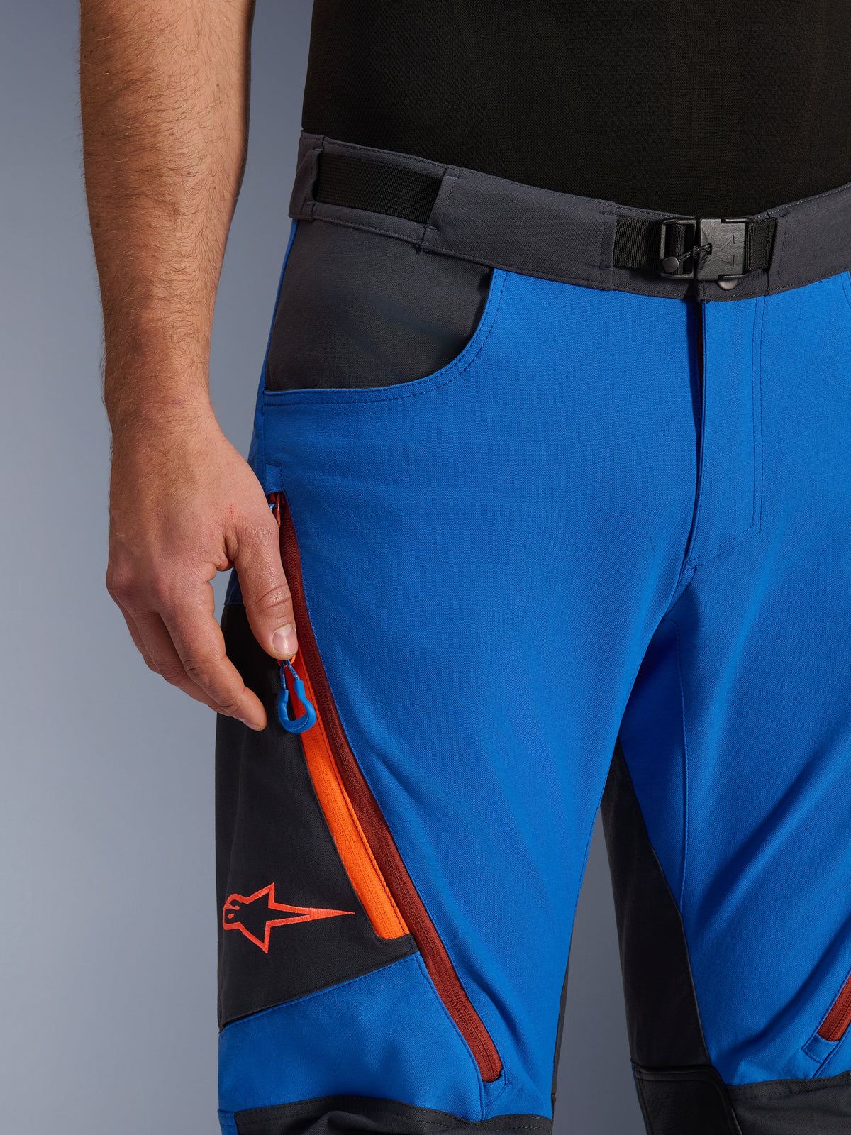 Alpinestars Maxdura Dual Hose in Royalblau und Dunkelgrau, Nahaufnahme des Hosenbundes mit Magnetverschluss, Cargotasche mit Reißverschluss, orangefarbenen Details und rotem Alpinestars-Logo, technische Adventure-Motorradausrüstung
