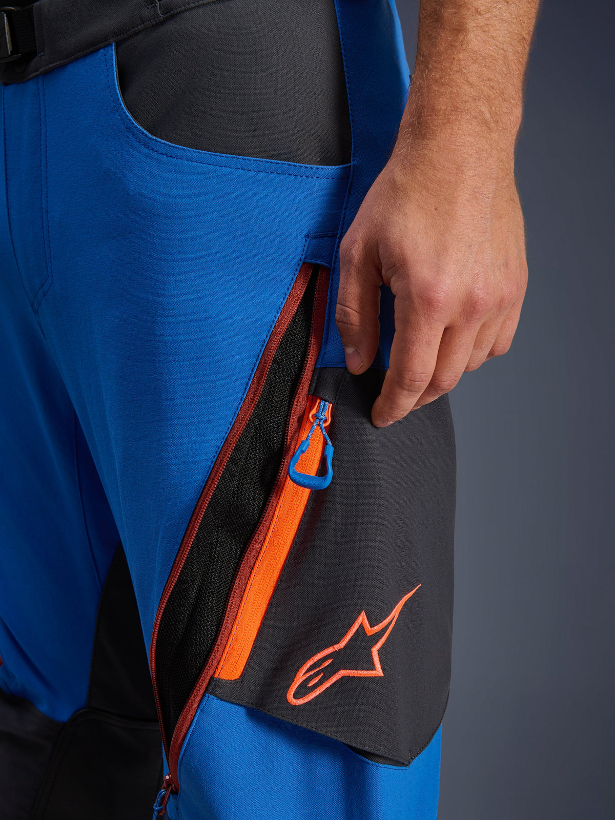 Alpinestars Maxdura Dual Hose Detail, Adventure-Motorradhose in Royalblau und Dunkelgrau, Nahaufnahme der Oberschenkelbelüftung mit Reißverschluss, orangefarbenen Akzenten und blauem Zugband, orangefarbenes Alpinestars-Logo auf schwarzem Panel, strapazierfähige Stretch-Polyamid-Konstruktion