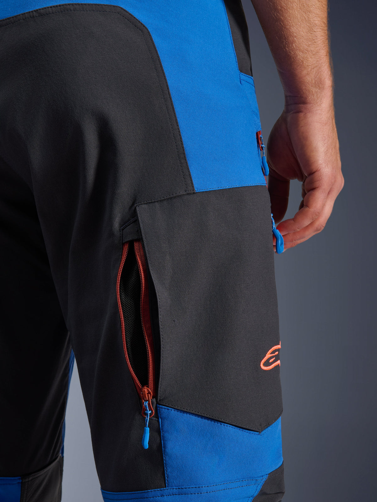 Alpinestars Maxdura Dual Hose, Königsblau und Dunkelgrau, Nahaufnahme der seitlichen Cargo-Tasche mit Reißverschluss, blaue Reißverschluss-Zipper, orangefarbenes gesticktes Logo, Adventure-Motorradbekleidung