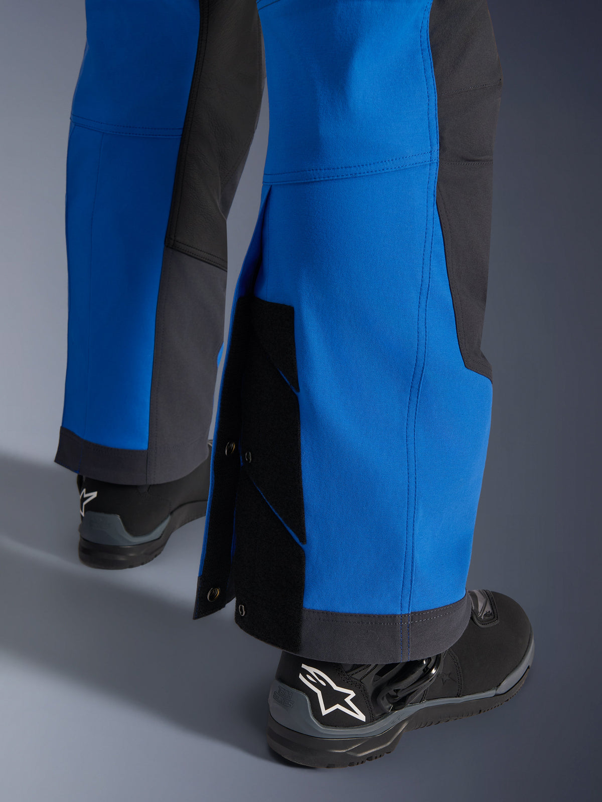 Alpinestars Maxdura Dual Hose, Königsblau und Dunkelgrau, Nahaufnahme des verstellbaren Hosenbeins mit Druckknopfverschlüssen und Zwickel, Über-den-Stiefel-Adventure-Passform, getragen mit schwarzen Alpinestars Motorradstiefeln