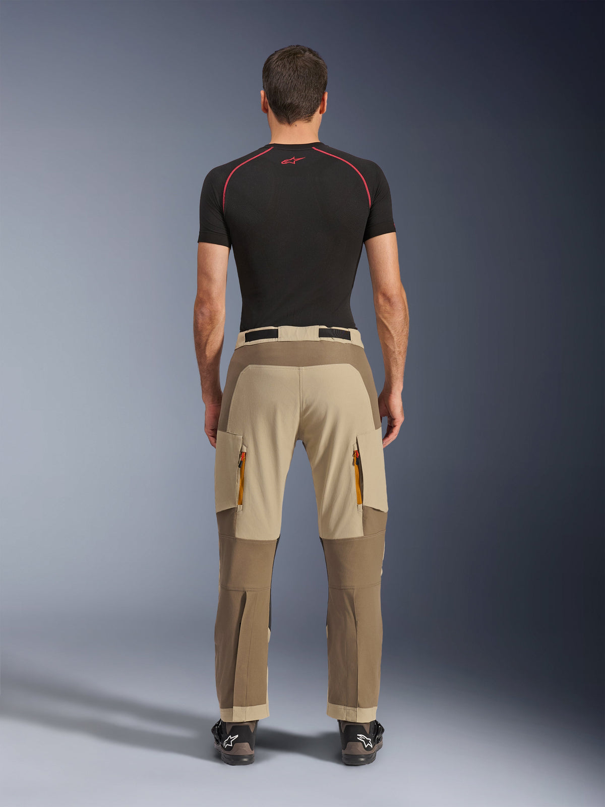 Alpinestars Maxdura Dual Hose in Walnut Aluminum, Rückansicht mit zweifarbigem Design in Braun und Tan, Adventure-Motorradhose mit Reißverschluss-Cargotaschen und verstärktem Gesäßbereich, getragen mit einem schwarzen Kompressionsshirt und Motorradstiefeln.