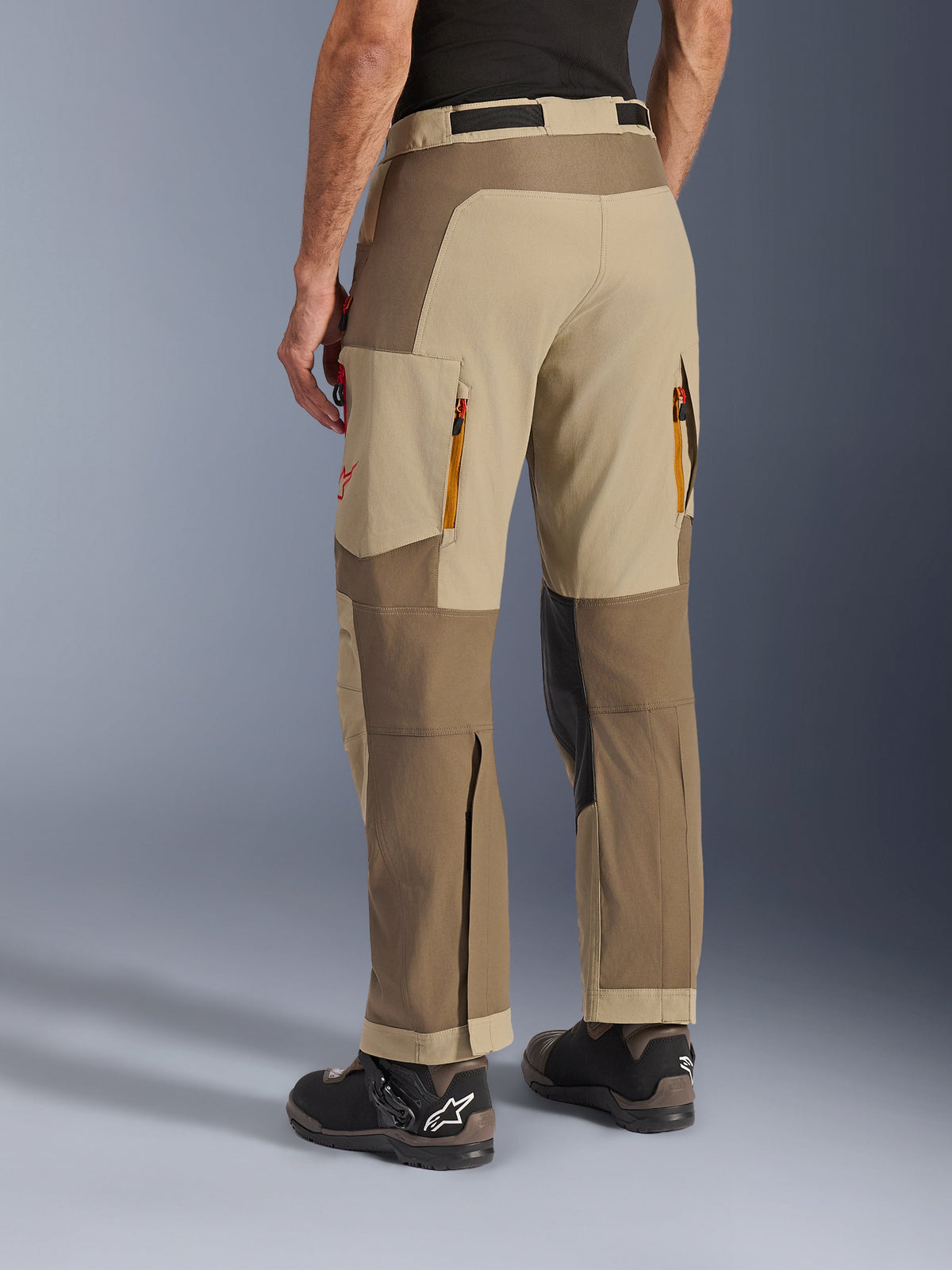 Alpinestars Maxdura Dual Hose in Walnut Aluminum, Rückansicht, Adventure-Motorradhose mit beige- und braunfarbenen Einsätzen, hintere Abluftöffnungen mit Reißverschluss, große Cargo-Taschen und Über-den-Stiefel-Design, getragen mit schwarz-braunen Stiefeln