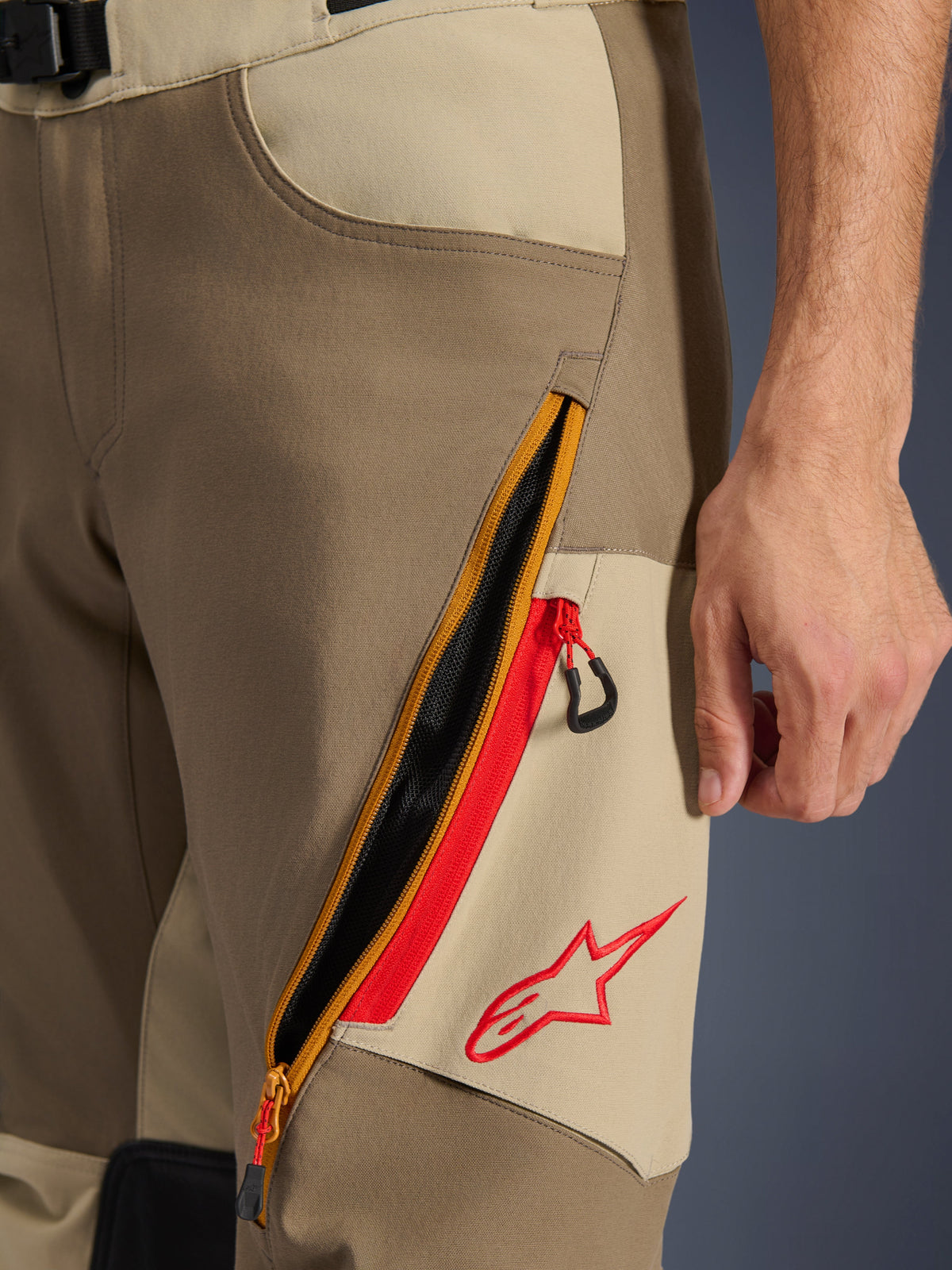 Alpinestars Maxdura Dual Hose, Walnuss und Aluminium, Nahaufnahme des Oberschenkel-Details mit großem diagonalen Reißverschluss-Belüftungssystem, rote Akzente und rotes Alpinestars-Logo auf beigem Einsatz, Adventure-Motorradausrüstung