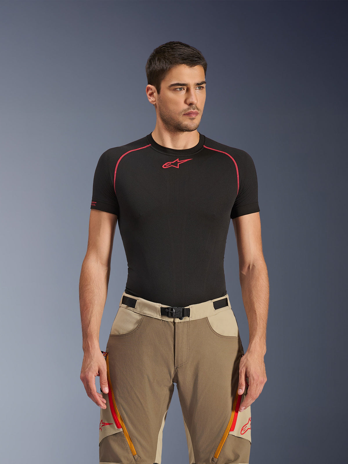 Alpinestars Maxdura Dual Hose in Walnut Aluminum, beige und sandfarbene Adventure-Motorradhose, getragen von einem Model mit schwarzem Base-Layer-Shirt, mit orangefarbenen Belüftungsreißverschlüssen an den Oberschenkeln, rotem Branding und einer magnetischen Taillenschnalle, Studioporträt.