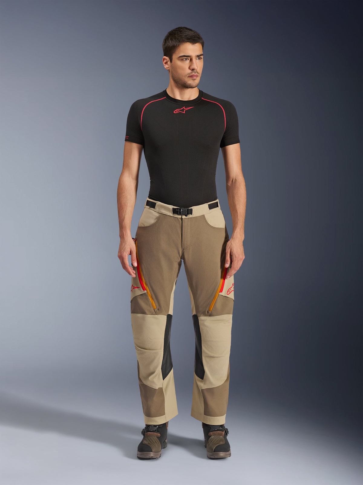 Alpinestars Maxdura Dual Hose, Adventure- und Dual-Sport-Motorradhose, Walnut Aluminum, getragen mit einem schwarzen kurzärmeligen Kompressionsshirt und Adventure-Stiefeln, mit orangefarbenen Belüftungsreißverschlüssen und roten Logos auf den Oberschenkeln, schwarzer Taillengürtel mit Schnalle, Über-den-Stiefel-Design zum Motorradfahren.