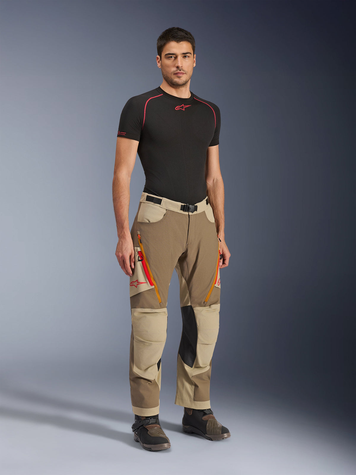Alpinestars Maxdura Dual Hose in Walnut Aluminum, braune und beigefarbene Adventure-Motorradhose mit orangefarbenen Reißverschlüssen und schwarzen Kniepaneelen aus Leder, getragen mit schwarzem Base-Layer-T-Shirt und Adventure-Stiefeln, Ganzkörper-Vorderansicht