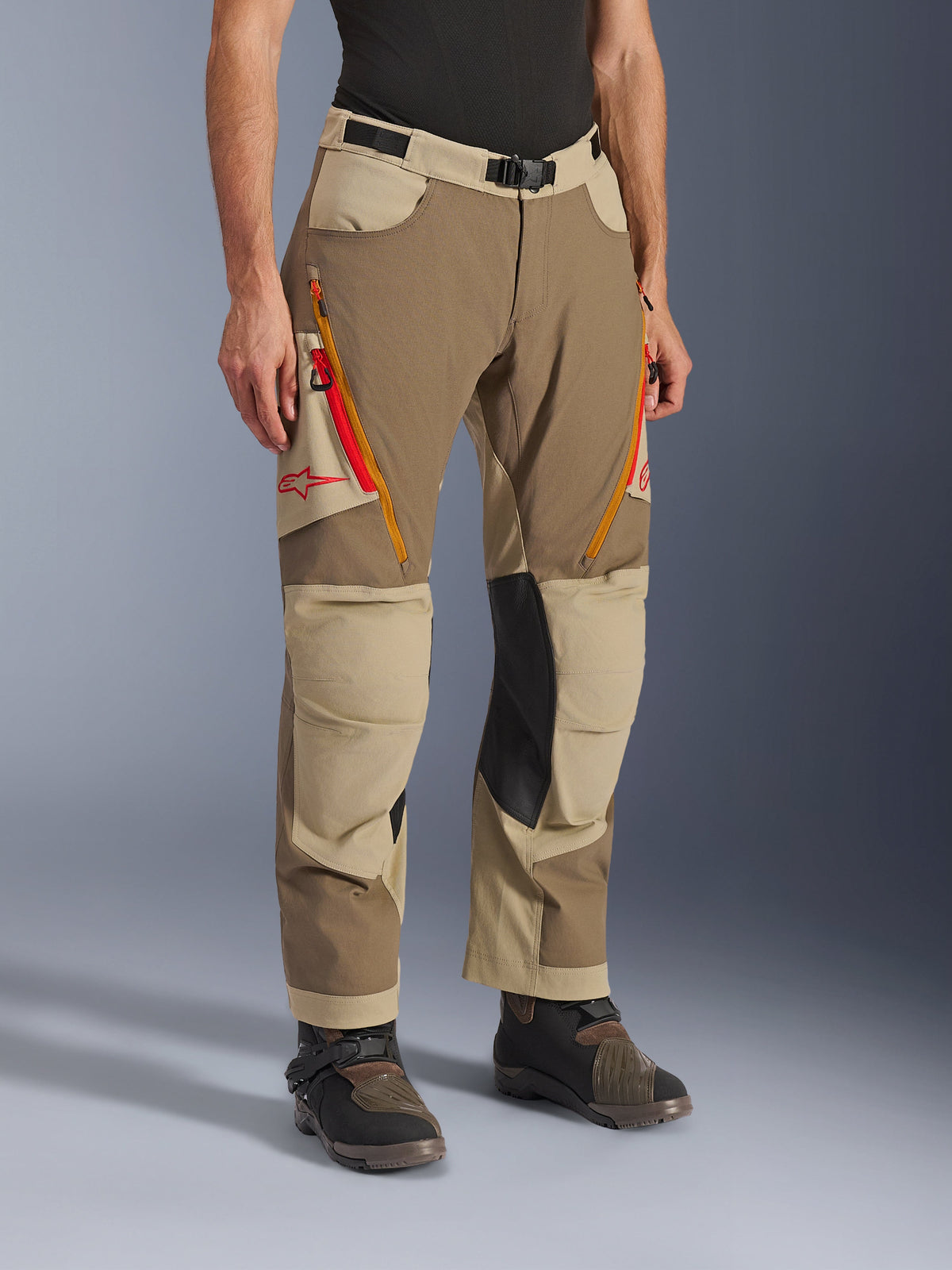Alpinestars Maxdura Dual Hose, walnut-aluminum-braune und khakifarbene Adventure-Fahrhose, mit orangefarbenen und roten Belüftungsreißverschlüssen an den Oberschenkeln, verstärkten schwarzen Leder-Innenknie-Einsätzen und rotem Astars-Logo, getragen mit technischen Stiefeln.