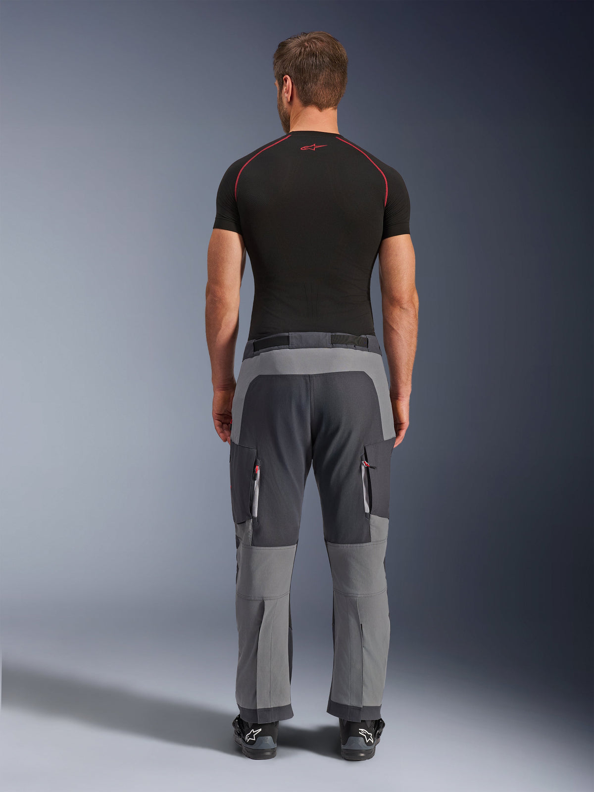 Alpinestars Maxdura Dual Hose, Adventure-Motorradhose in Dunkelgrau und Deep Gray, Rückansicht am Model in schwarzem T-Shirt und Stiefeln, mit mehrfarbigen grauen Einsätzen und Reißverschluss-Cargotaschen mit roten Akzenten.