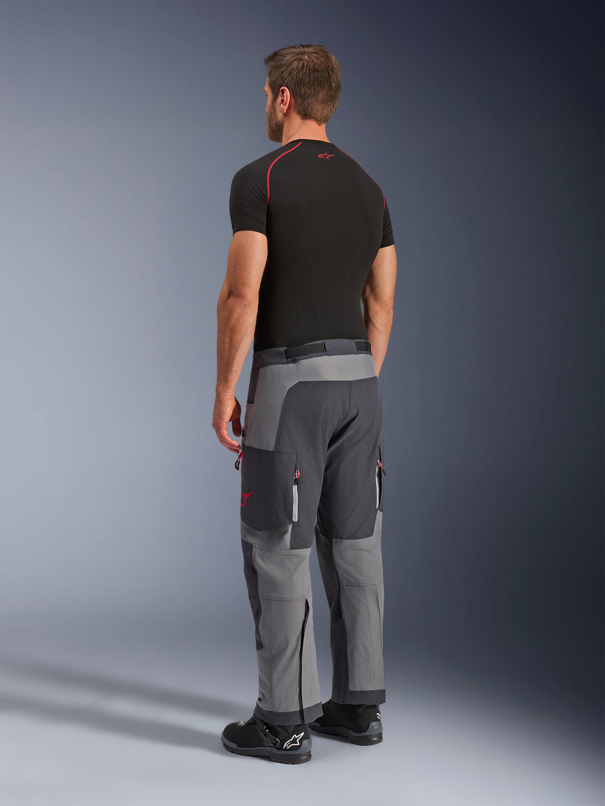 Alpinestars Maxdura Dual Hose, Adventure-Motorradhose, Dunkelgrau und Tiefgrau, Rückansicht am Modell, rote Akzente an Reißverschlüssen und Logo, verstärkte graue Paneele, Cargotaschen und Over-Boot-Passform