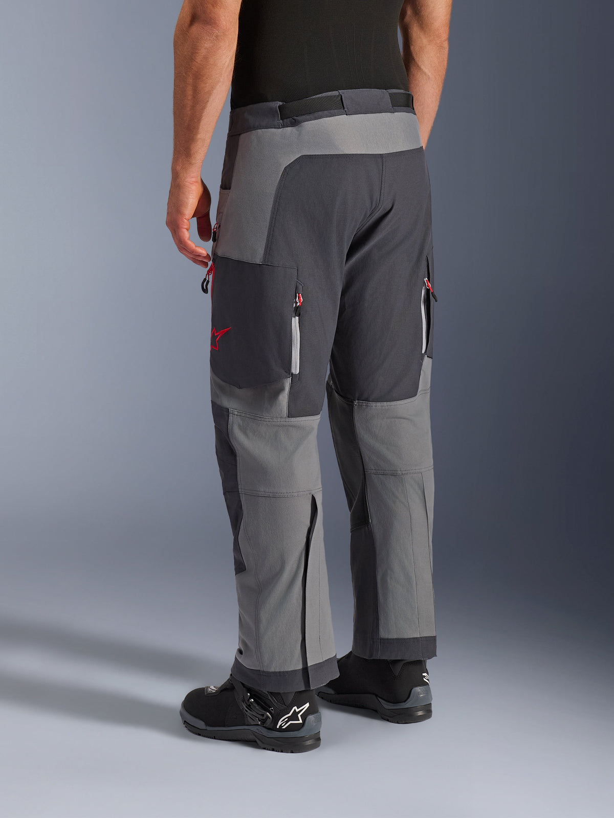 Alpinestars Maxdura Dual Hose, Adventure-Motorradhose, Dunkelgrau und Tiefgrau, Rückansicht mit Multi-Panel-Konstruktion, roten Reißverschluss-Akzenten und Stern-Logo, kombiniert mit schwarzen Alpinestars-Stiefeln, entwickelt für Dual-Sport-Performance