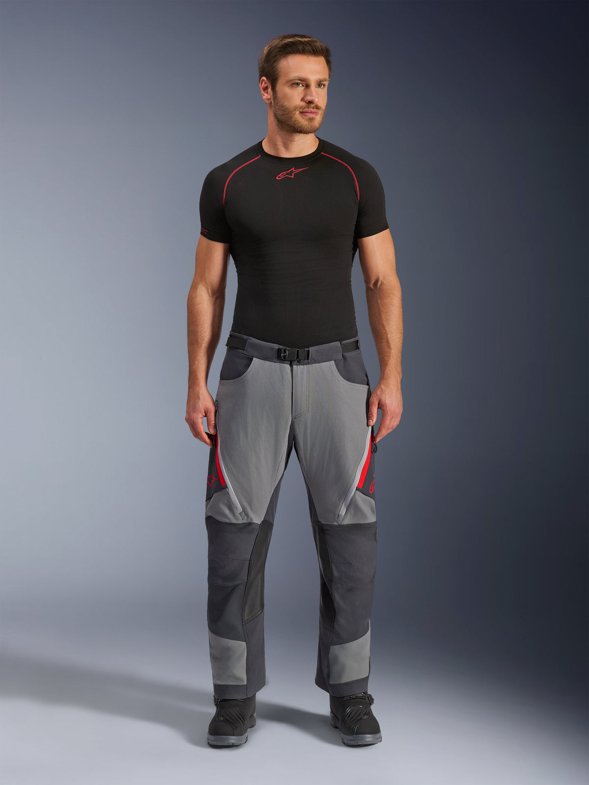 Alpinestars Maxdura Dual Hose, Motorradhose, Dunkelgrau und Tiefgrau mit roten Akzenten, Vorderansicht getragen von einem Model mit schwarzem Alpinestars Base Layer und schwarzen Stiefeln, mit verstärkten Paneelen und Oberschenkel-Belüftungsöffnungen mit Reißverschluss, Adventure-Stil Fahrausrüstung