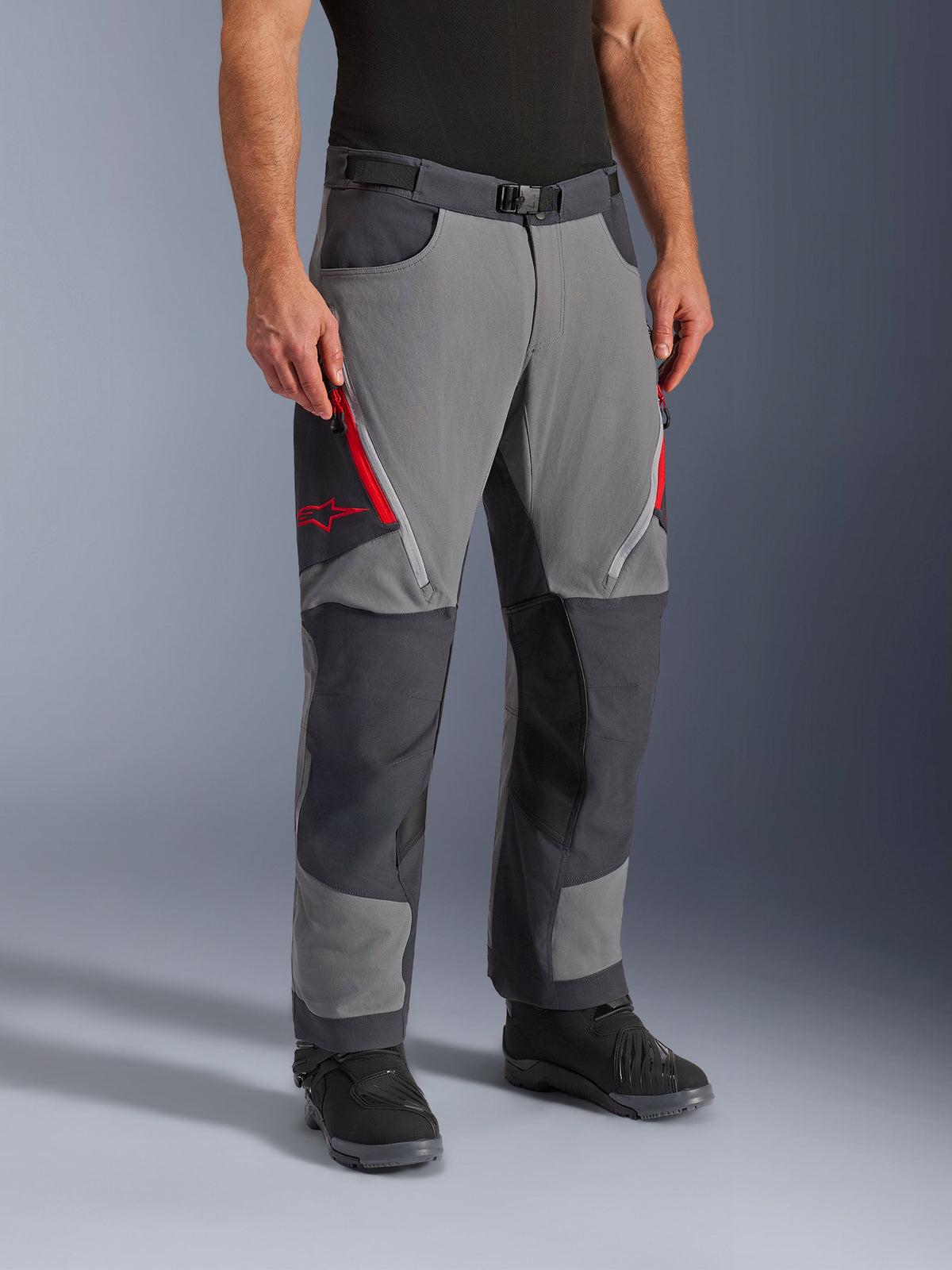 Alpinestars Maxdura Dual Hose in Dunkelgrau und Tiefgrau, Adventure-Touring-Motorradhose mit roten Belüftungsreißverschlüssen an den Oberschenkeln und Stern-Logo, verstärkte dunkle Kniepaneele, kombiniert mit schwarzen Motorradstiefeln