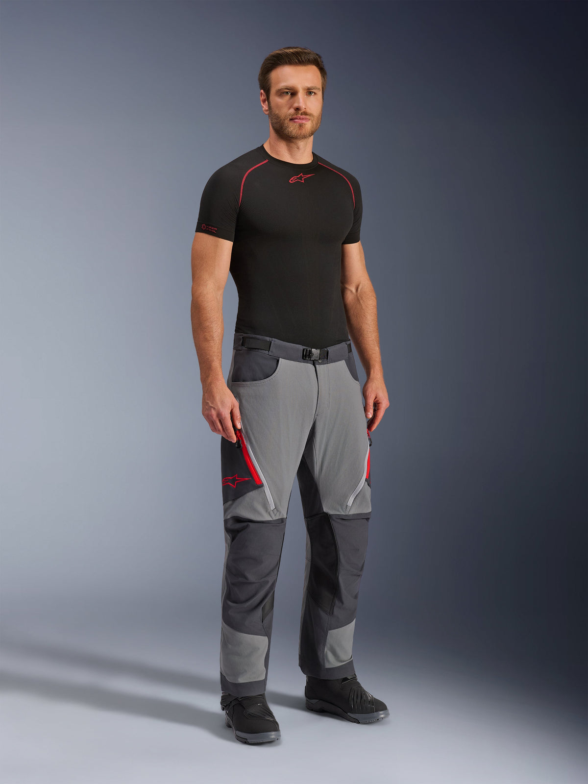 Alpinestars Maxdura Dual Hose, Adventure-Motorradhose, Dunkelgrau und Tiefgrau, getragen mit schwarzem Base Layer und Stiefeln, mit roten Belüftungsreißverschlüssen, verstärkten Einsätzen an Knien und Gesäß sowie einem Taillengürtel, sportliche Passform für den Dual-Sport-Einsatz