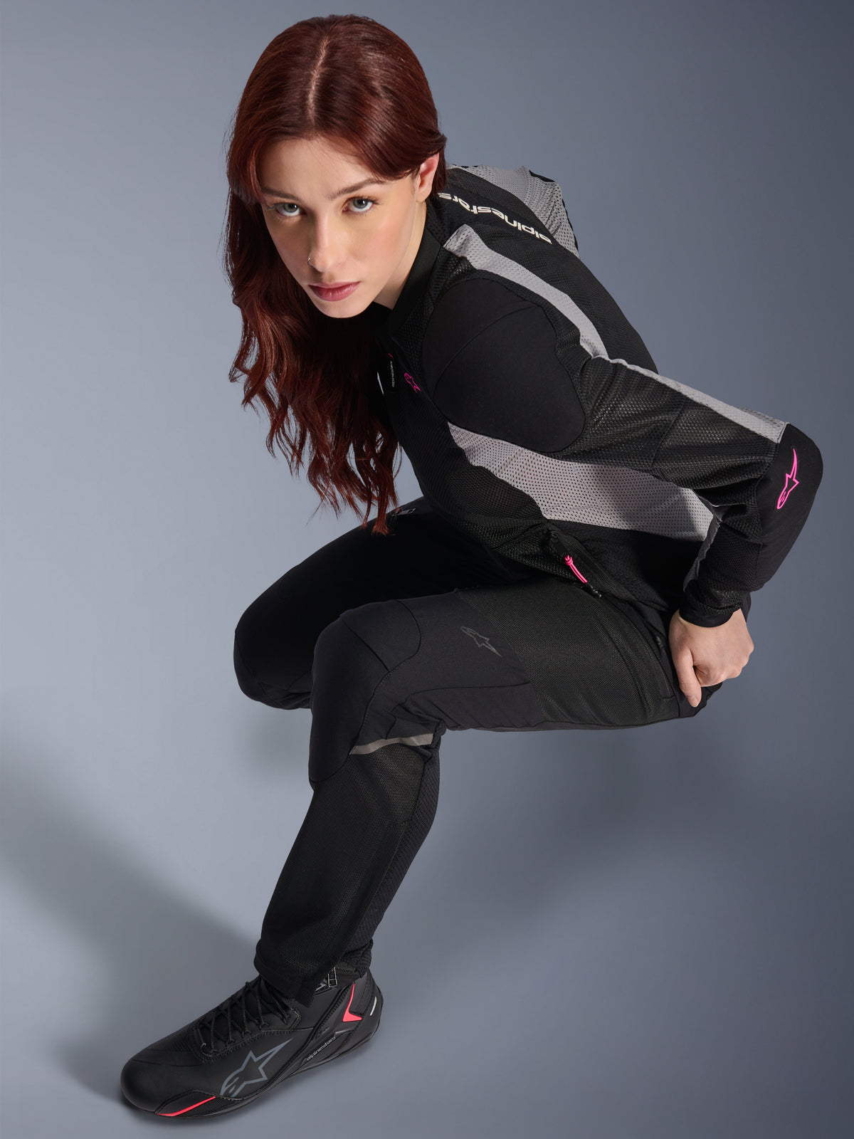 Frau trägt Alpinestars Stella Troop-Air Hose, Schwarz, leichte belüftete Textilhose, kombiniert mit einer schwarz-grauen Mesh-Jacke mit rosa Akzenten und schwarzen Motorradstiefeln, Studioporträt, urbane Motorradbekleidung